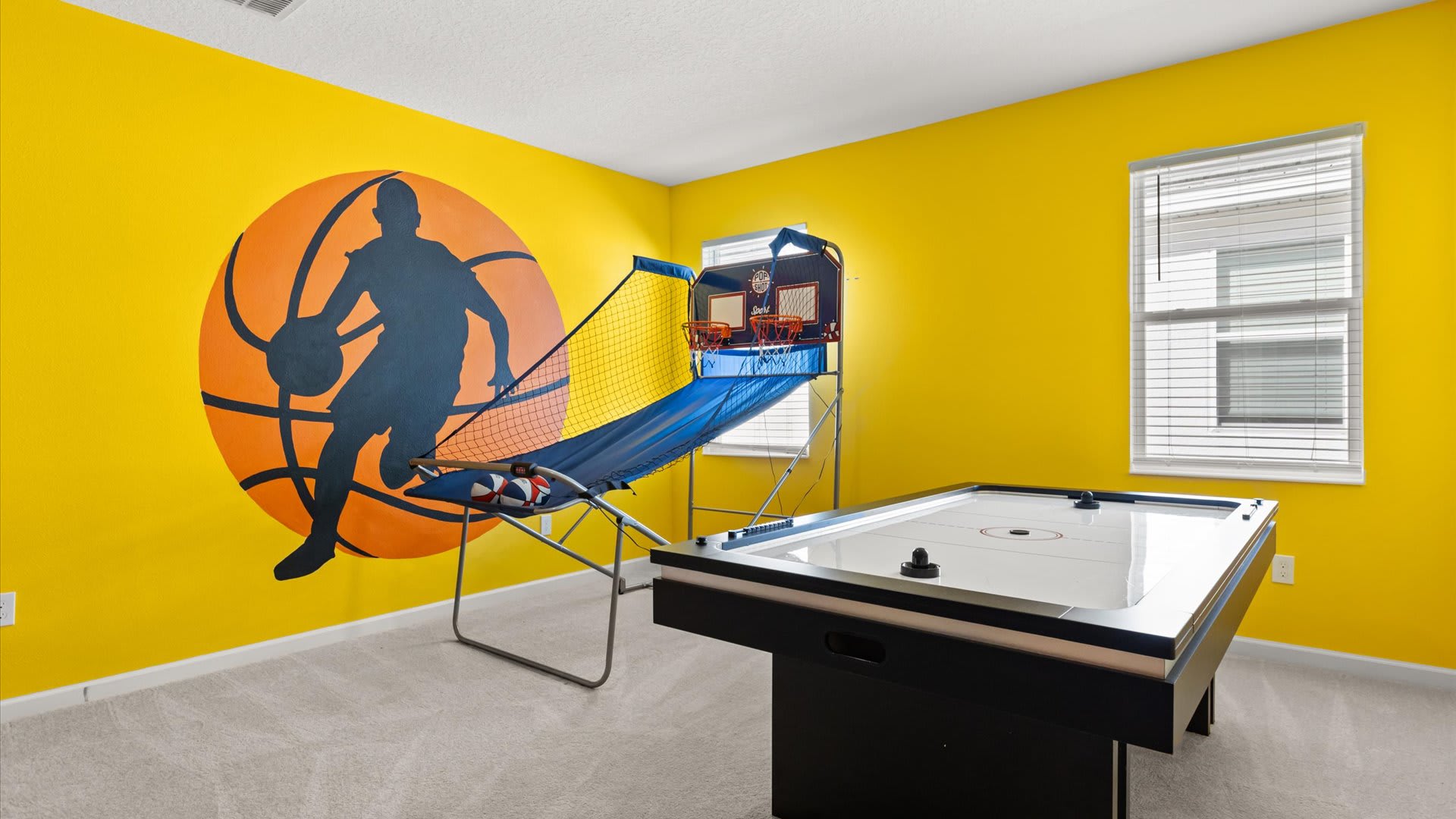 Vibrant game room with bright yellow walls, one featuring a large basketball player mural. The room includes a basketball arcade game and an air hockey table, with carpeted flooring.