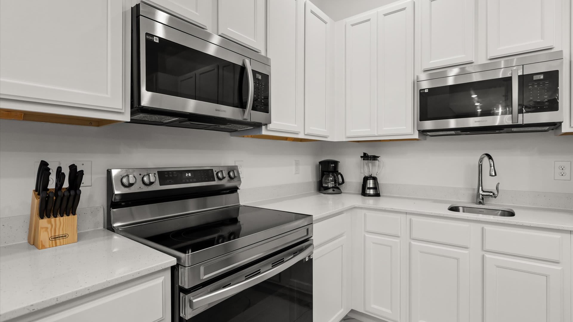 Detailed view of the kitchen counter with white shaker cabinets, a stainless steel electric range, microwave, coffee maker, and blender. The white quartz countertop provides ample prep space.