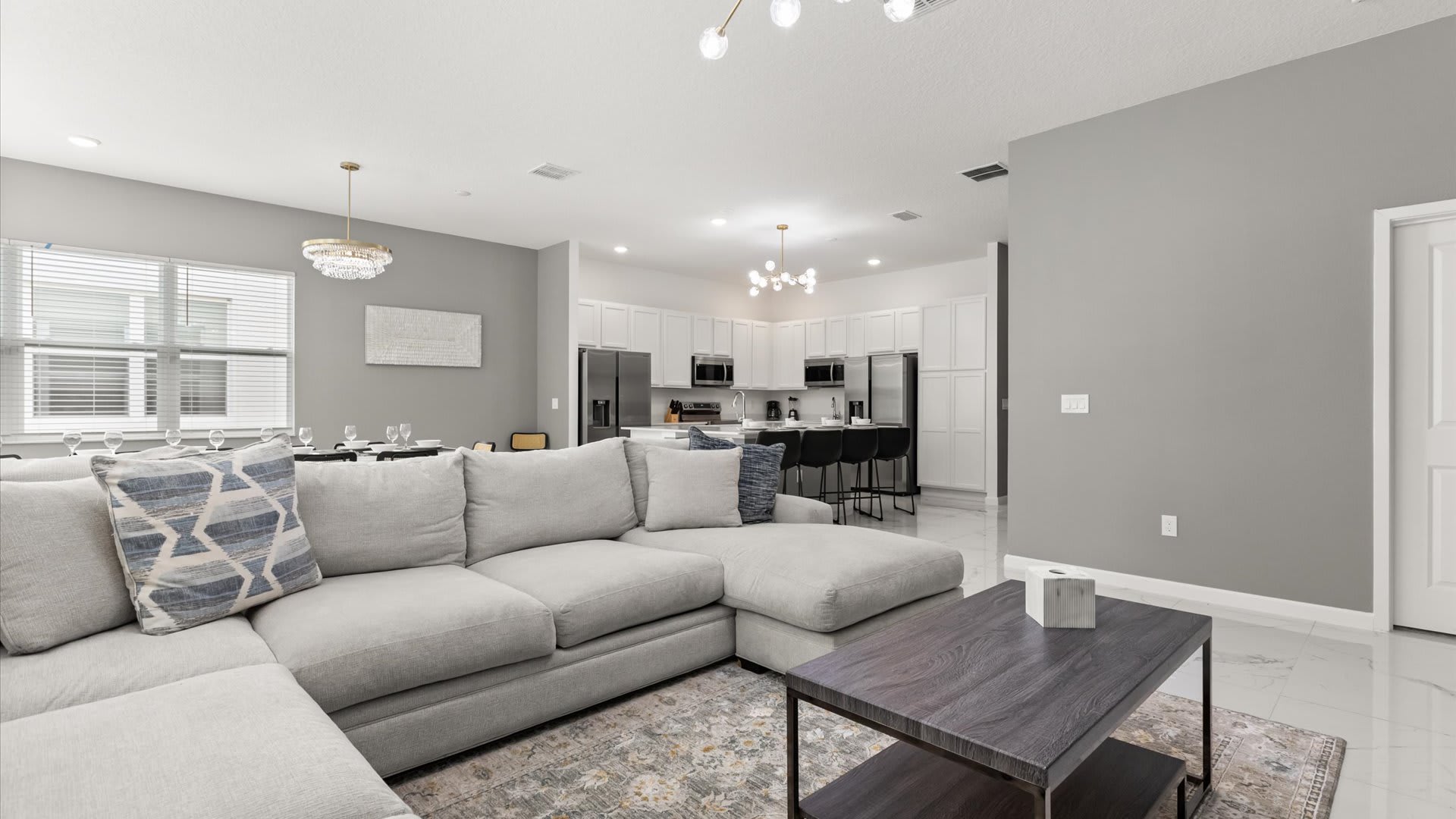 An expansive open-concept living area features a large light gray sectional sofa and a dark wood coffee table on a patterned rug. The space flows into a modern kitchen with white cabinetry and a breakfast bar, with a dining area visible in the background.