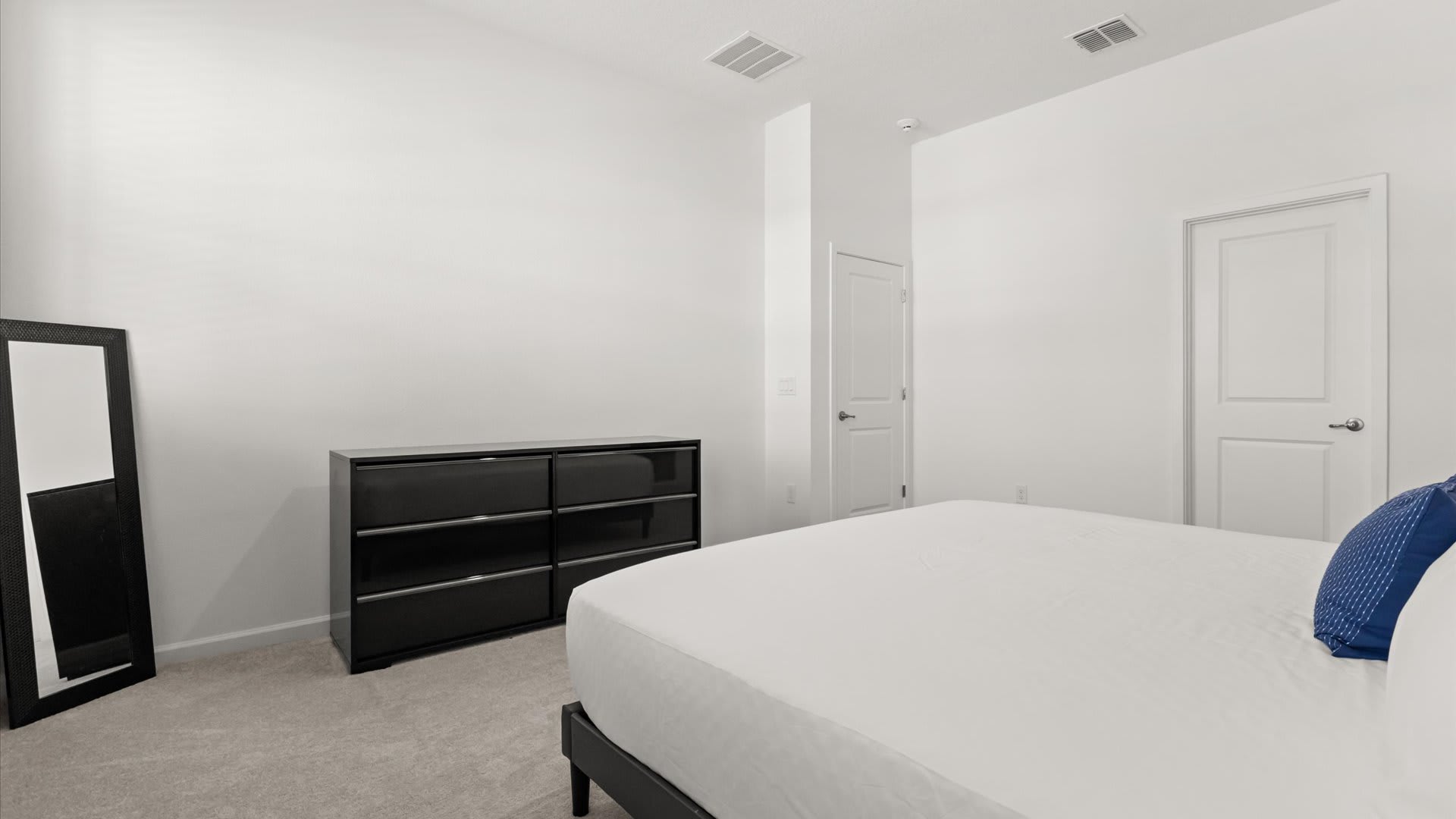 A bright bedroom with white walls, featuring a dark wood dresser with silver accents and a tall black-framed full-length mirror leaning against the wall. A bed with white bedding and a blue accent pillow is visible on the right.