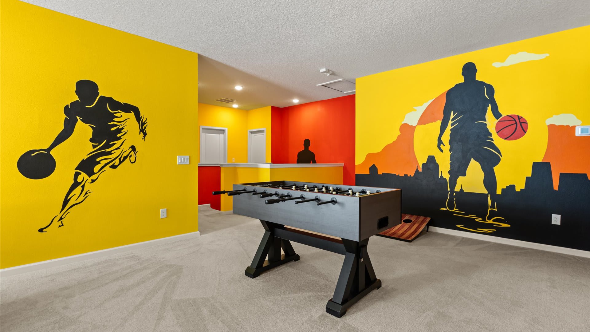A vibrant game room with bright yellow and red walls adorned with large black silhouette murals of basketball players. A foosball table is centrally located on the light gray carpet, with a cornhole board nearby.