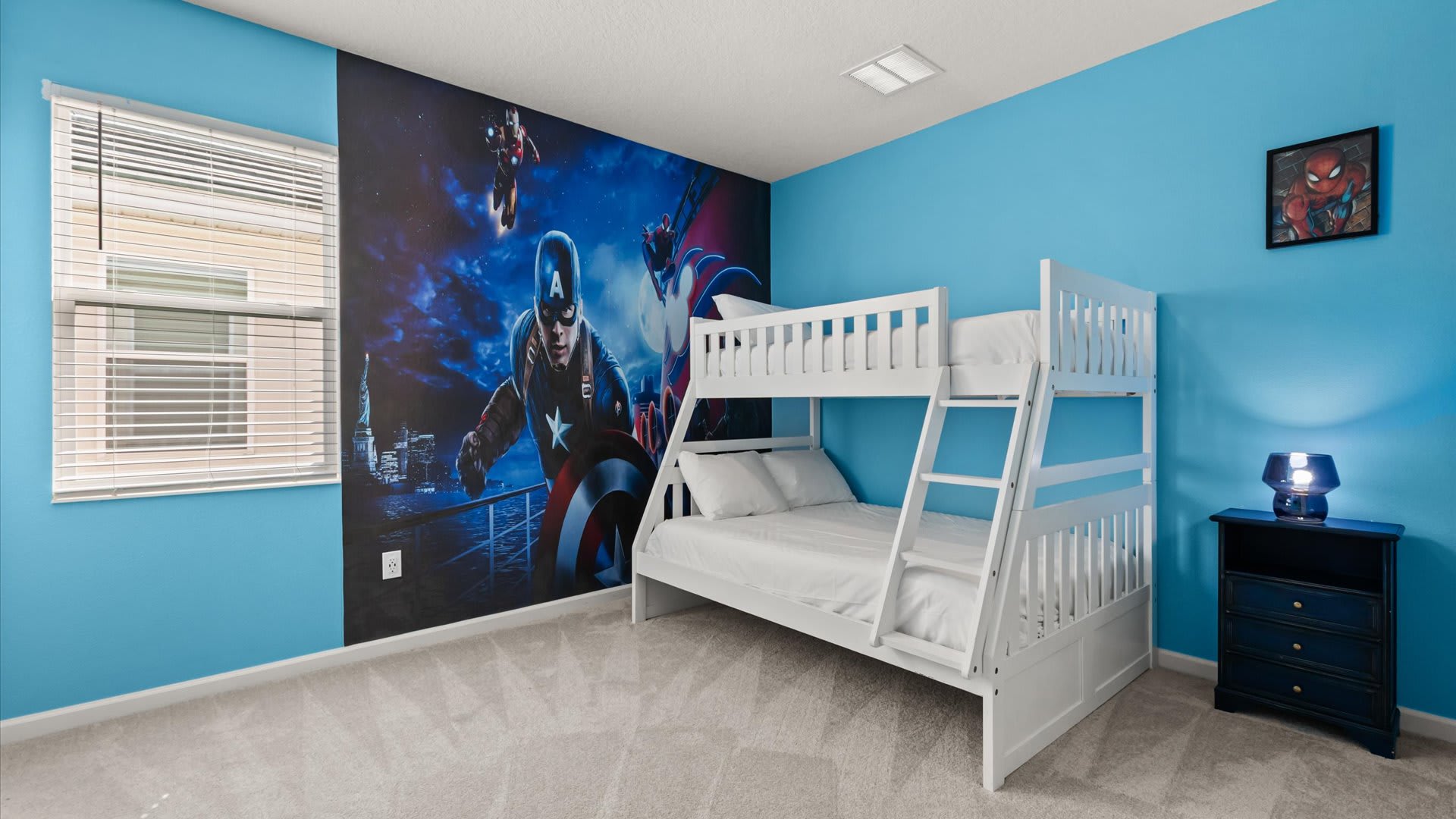 A themed kids' bedroom features a white wooden bunk bed with white bedding against a striking superhero mural wall. The room has bright blue walls and a small dark blue nightstand with a lamp.