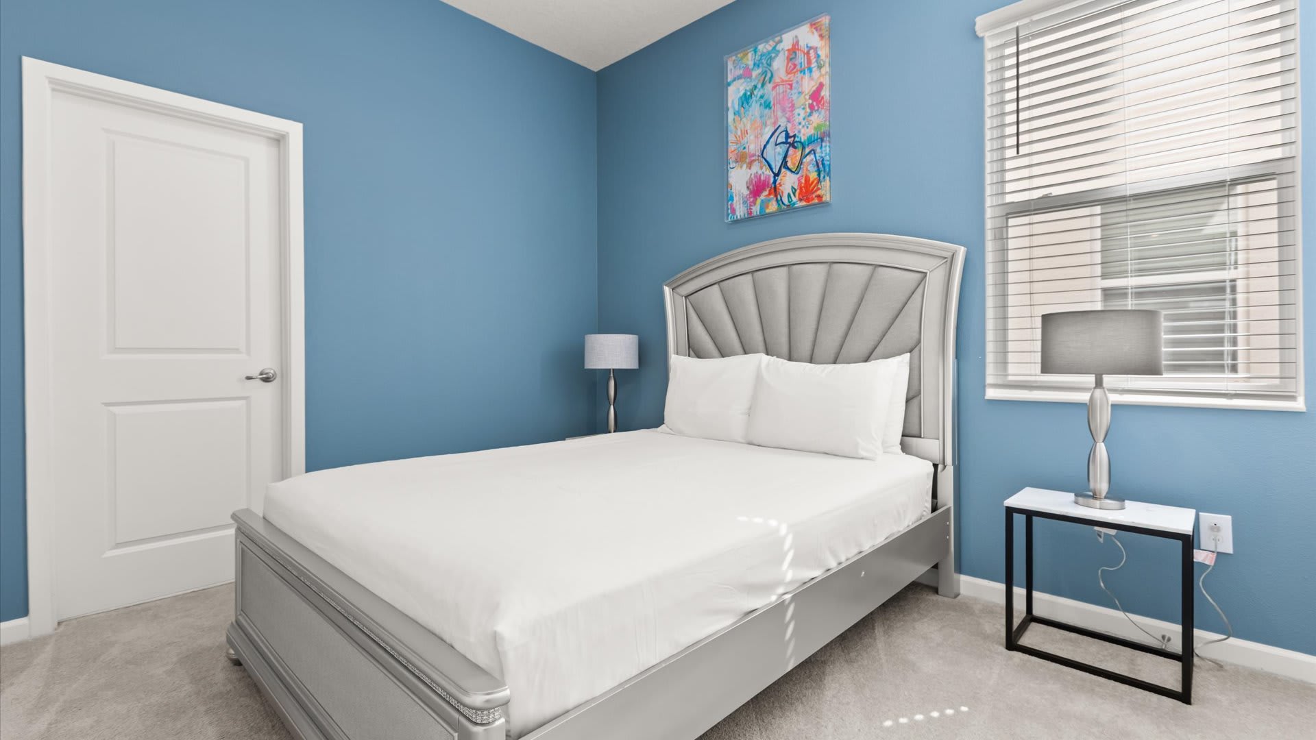 A vibrant bedroom with blue walls features a silver-colored bed frame with a padded headboard and white bedding. A small black nightstand with a lamp is beside the bed, and colorful abstract art hangs above the headboard.
