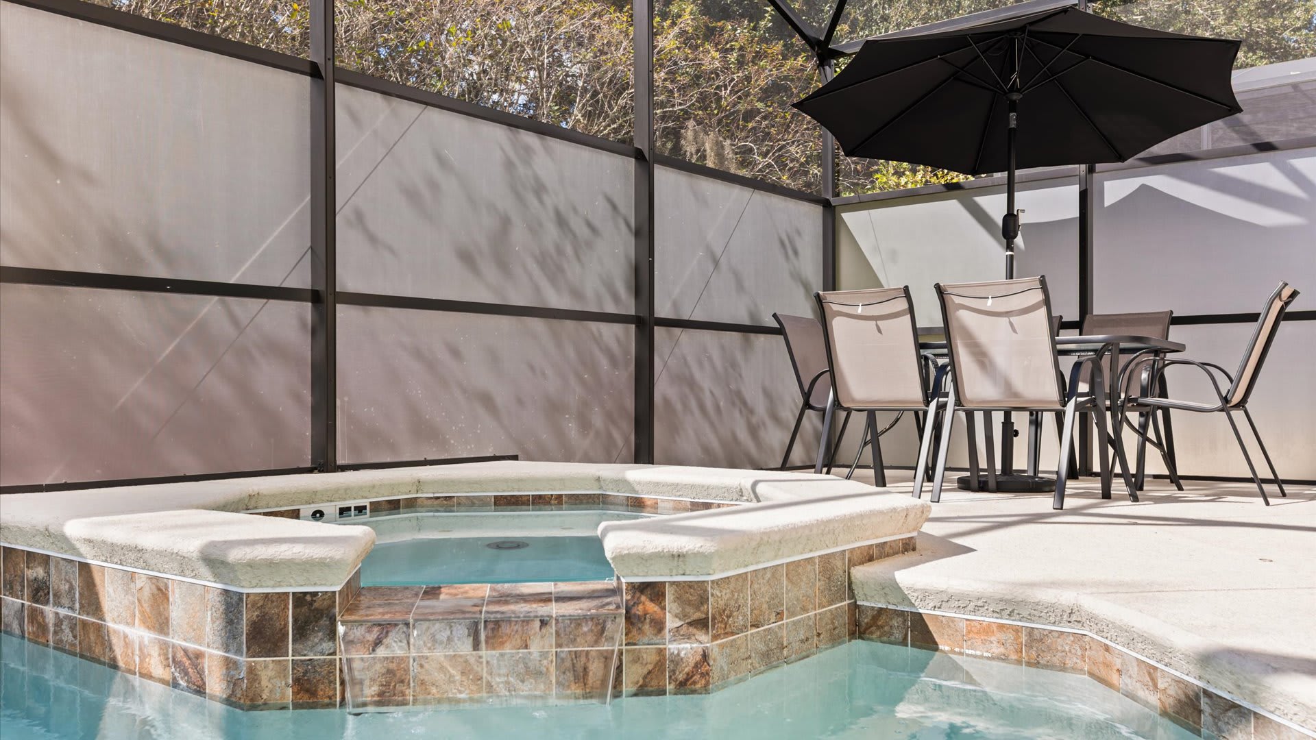 A close-up view of the tiled hot tub within a screened enclosure, adjacent to an outdoor dining area with a table and chairs shaded by a black umbrella. Opaque privacy screens surround the area.