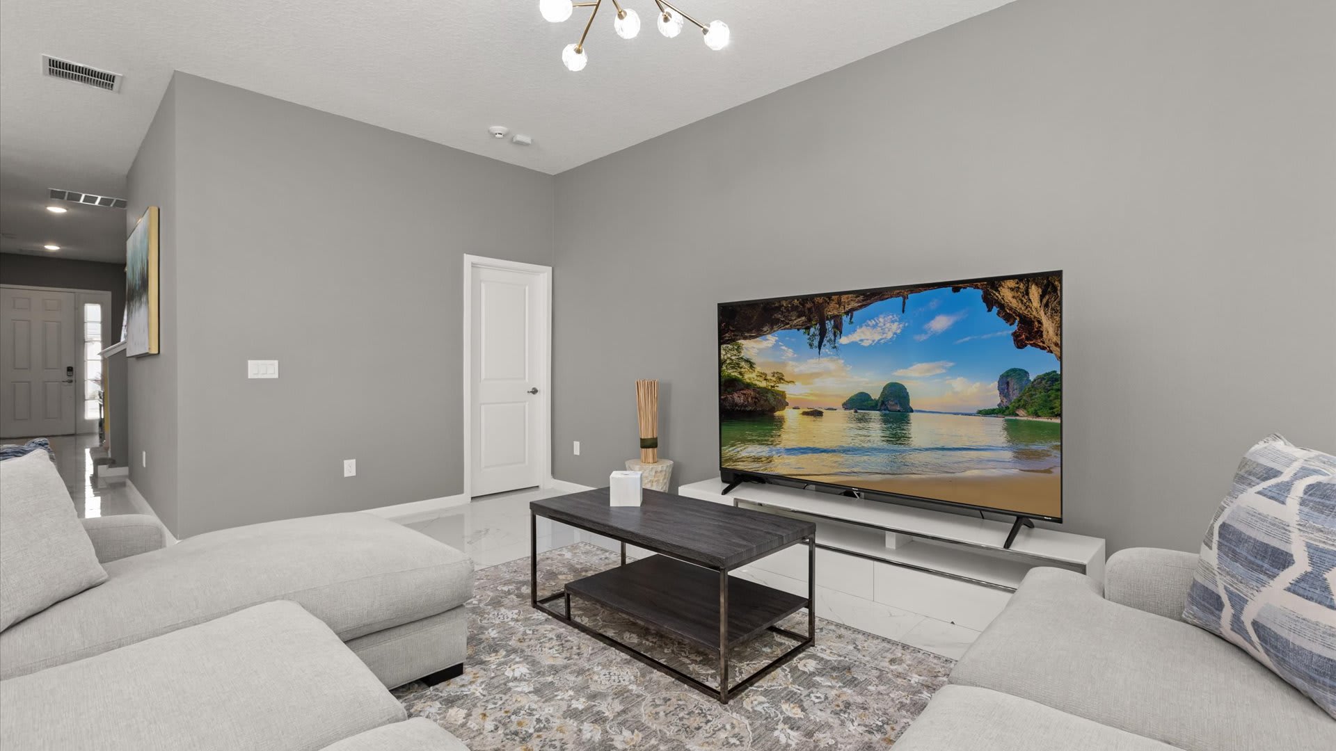 Modern living room with light gray walls, a large L-shaped sectional sofa, a dark wood coffee table, and a big screen TV displaying a beach scene. A contemporary chandelier hangs from the ceiling.