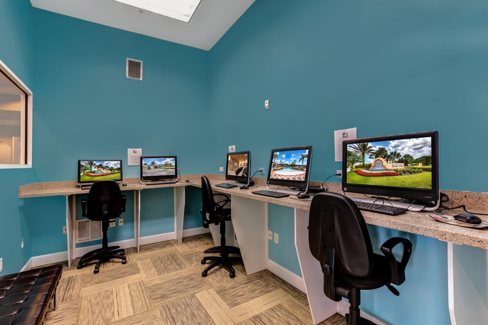 A communal business center with light blue walls and patterned carpet, featuring multiple computer workstations with desktop computers and office chairs. A long counter provides ample workspace, and a window offers a view to an adjacent room.