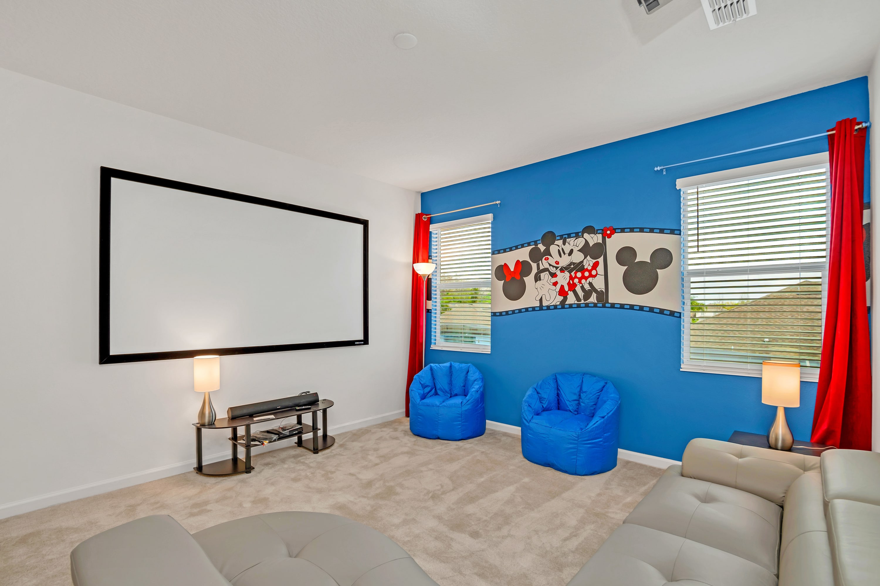 Dedicated media room featuring a large projection screen, a comfortable gray sectional sofa, two vibrant blue bean bag chairs, and a playful blue accent wall adorned with Mickey and Minnie Mouse decor.