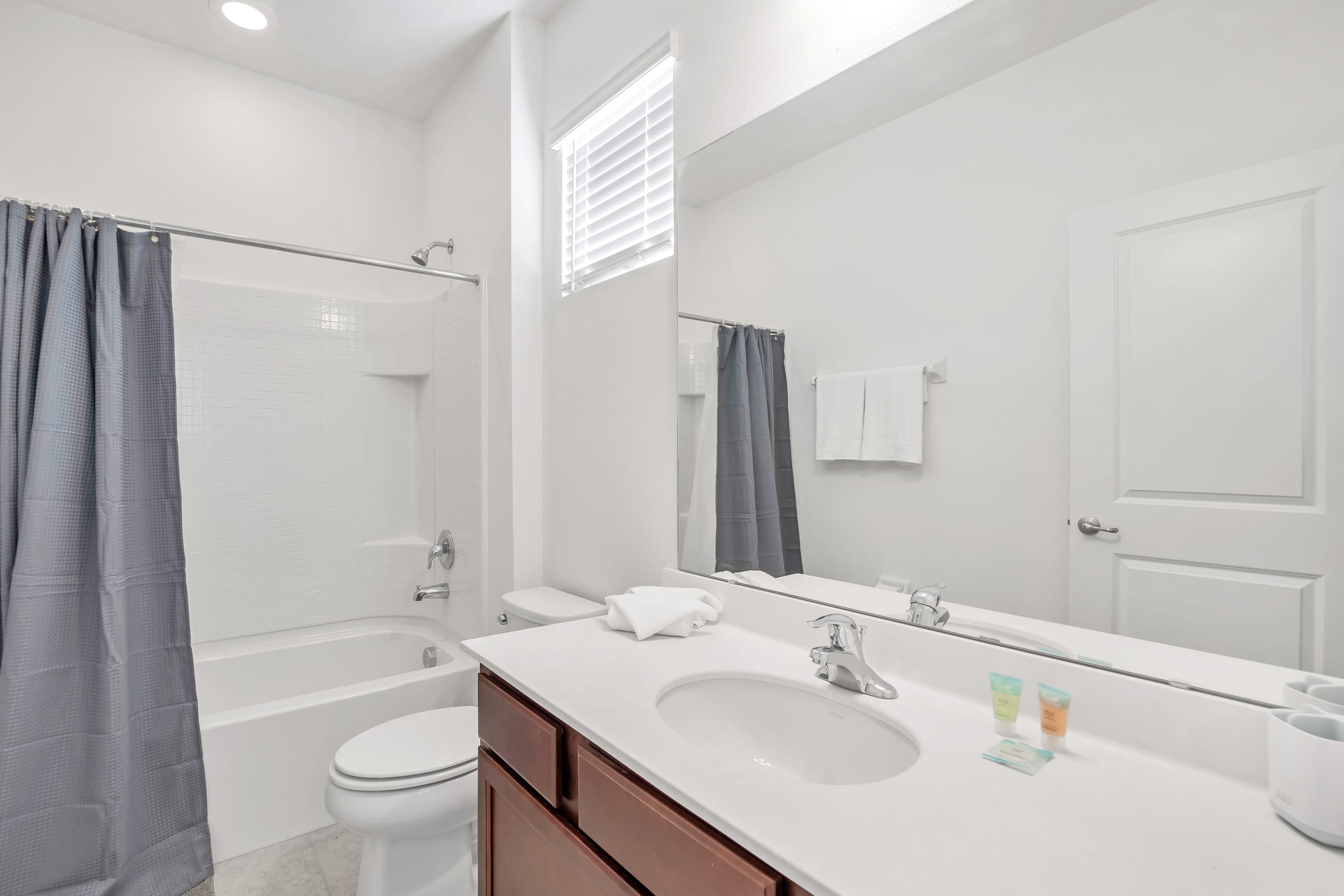 A clean white bathroom featuring a dark wood vanity with a single sink, a large mirror, and a toilet. A white tiled shower/tub combination with a dark blue shower curtain is visible on the left.