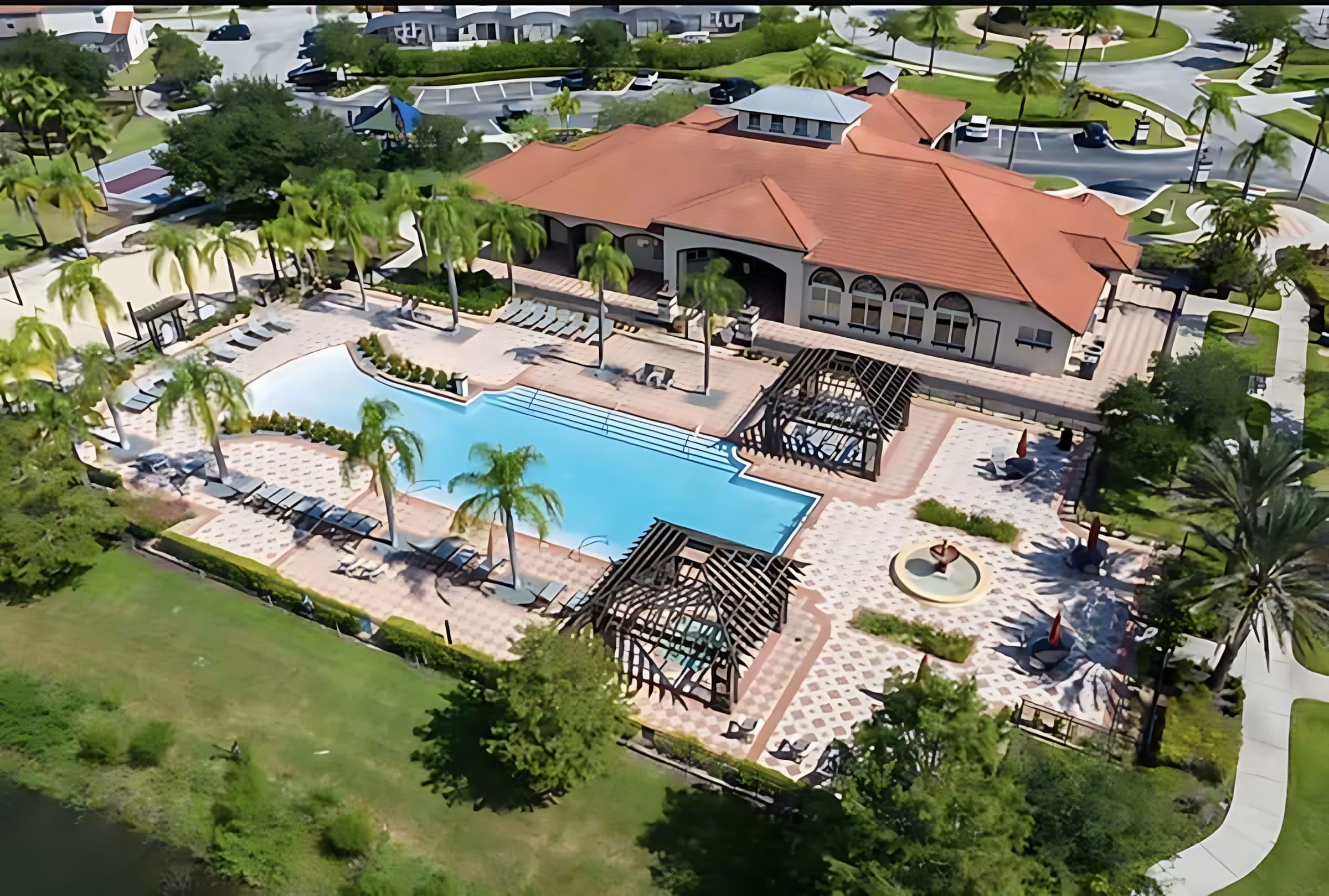 An aerial view of a large communal swimming pool area with lounge chairs, pergolas, and palm trees. A large clubhouse building with a red tile roof is visible in the background, surrounded by green landscaping.