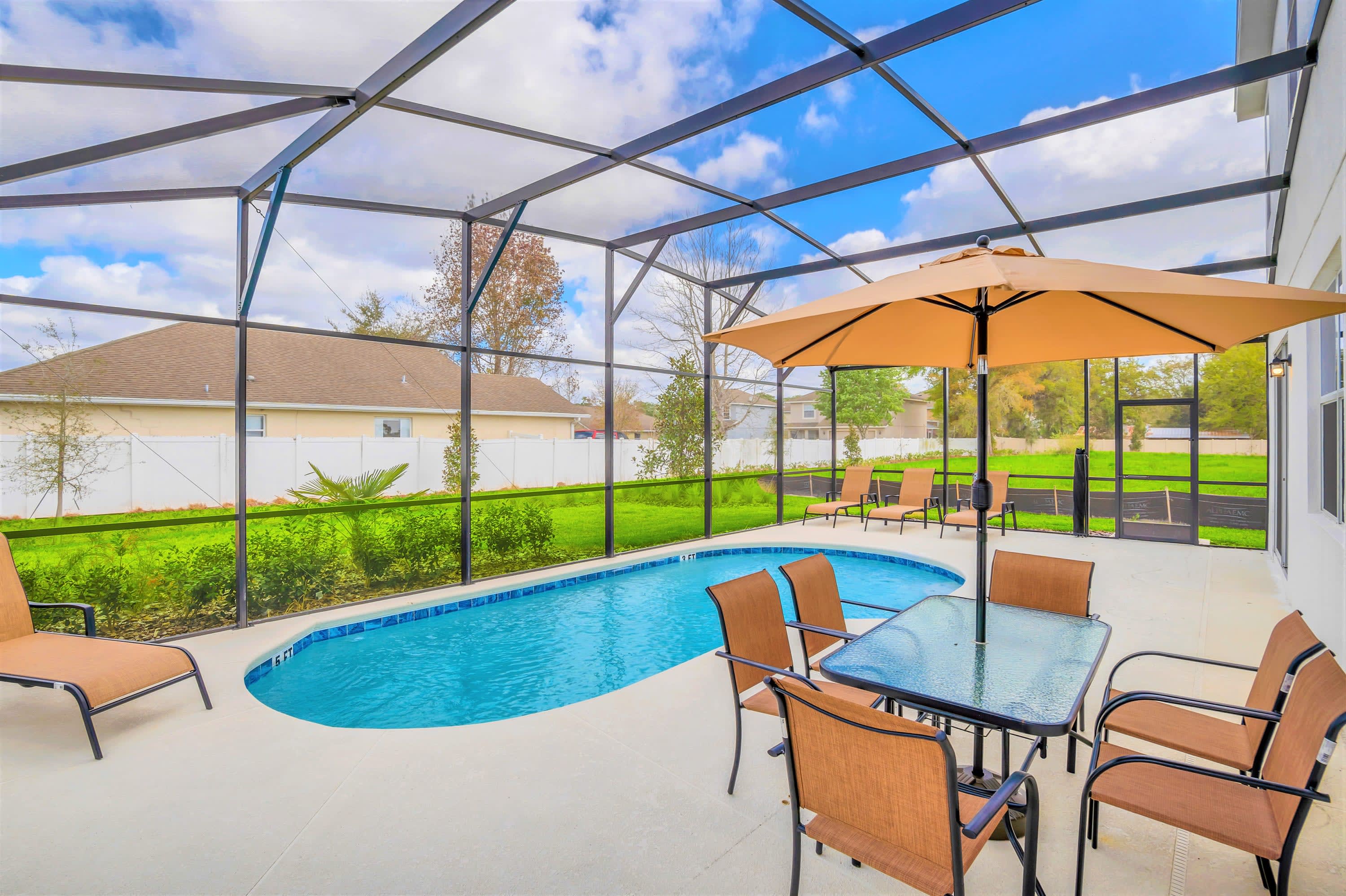Screened-in private pool area featuring a kidney-shaped pool, an outdoor dining table with an umbrella, and several lounge chairs on a light-colored concrete deck. The enclosure offers views of a grassy backyard and neighboring houses under a bright blue sky.