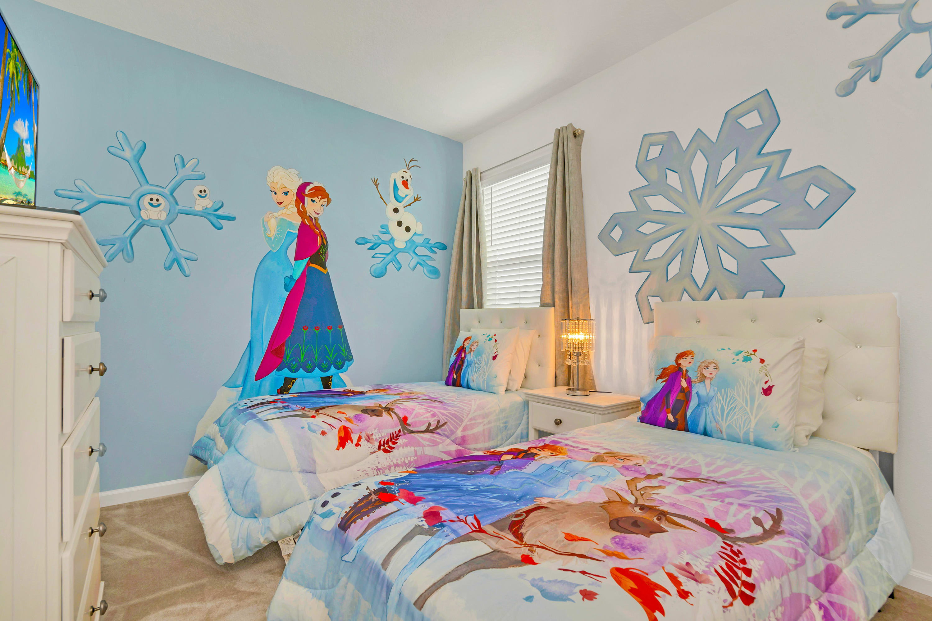 A whimsical children's bedroom featuring two twin beds with colorful Frozen-themed bedding and large wall decals of Elsa, Anna, and Olaf. A white dresser and nightstand with a lamp are also present.