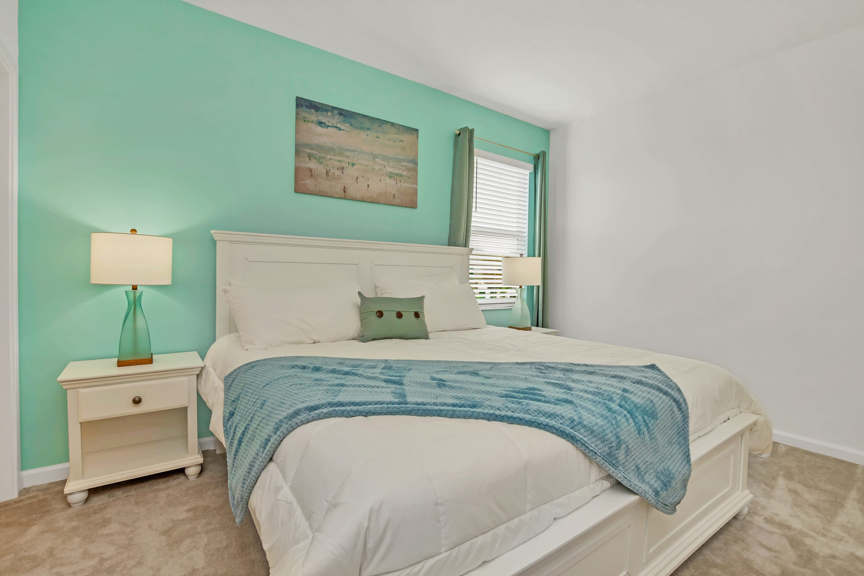 A vibrant bedroom with a white king-sized bed, a teal throw blanket, and two white nightstands with lamps. A teal accent wall and a framed beach artwork create a refreshing, coastal-inspired atmosphere.