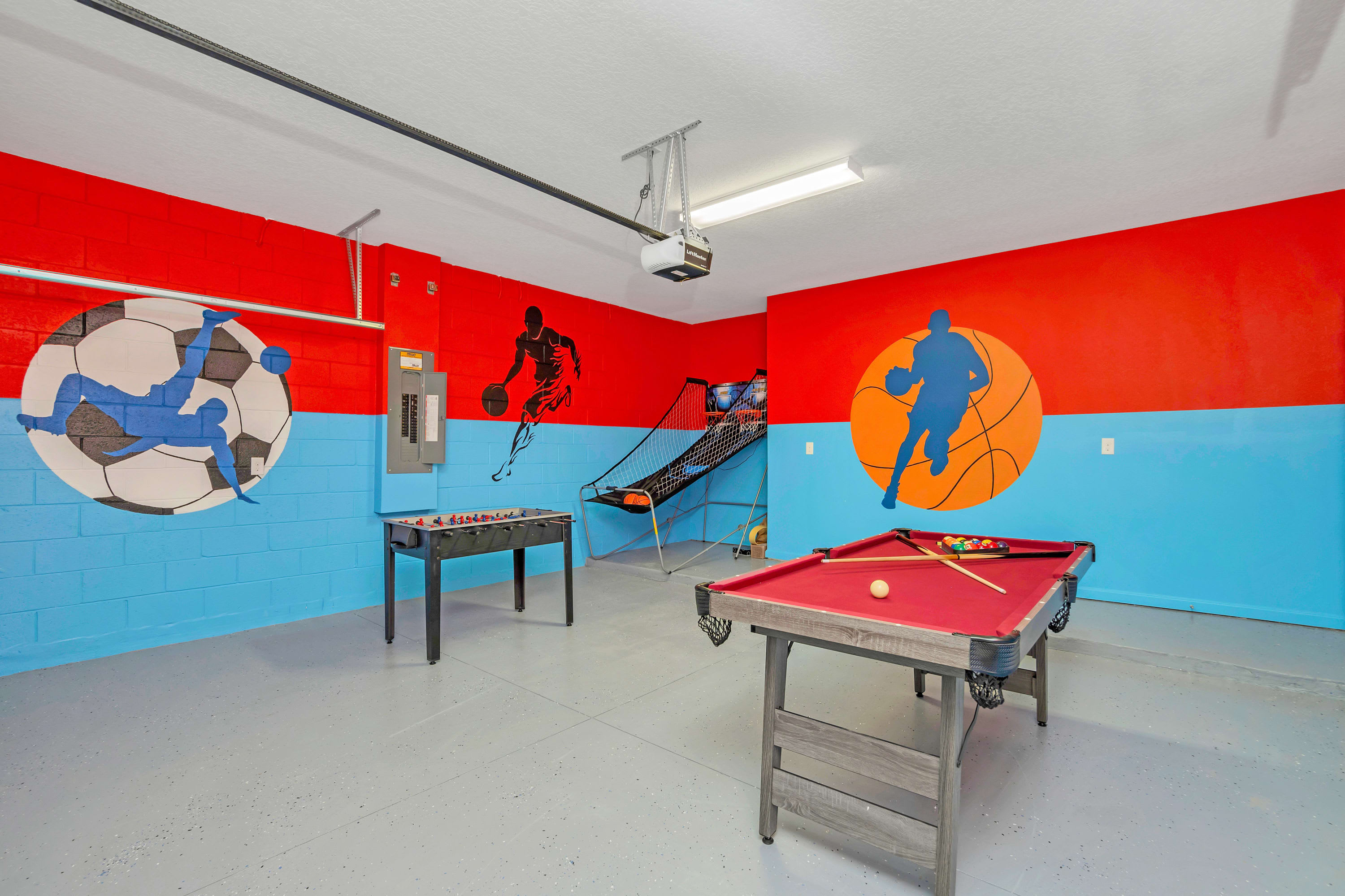 Spacious game room with red and blue walls adorned with sports-themed murals, featuring a foosball table, a basketball arcade game, and a red felt pool table.
