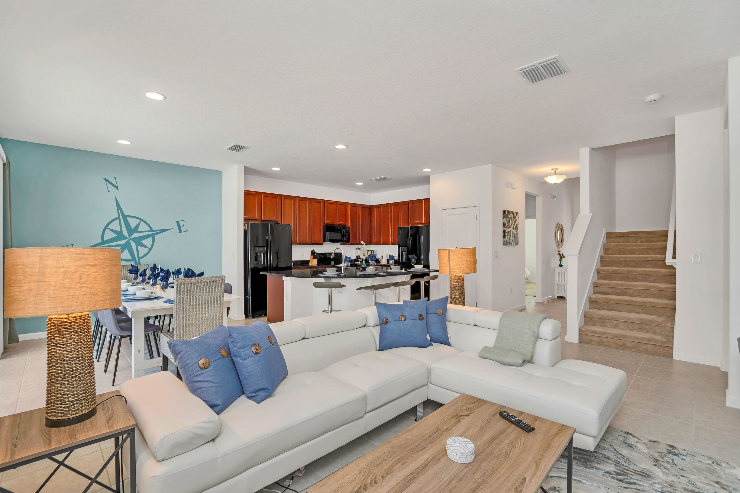 Open-concept living space with a large white sectional sofa, wooden coffee table, and a view of the dining area with a teal accent wall and the kitchen featuring dark wood cabinets and black appliances. Stairs lead to the upper floor.