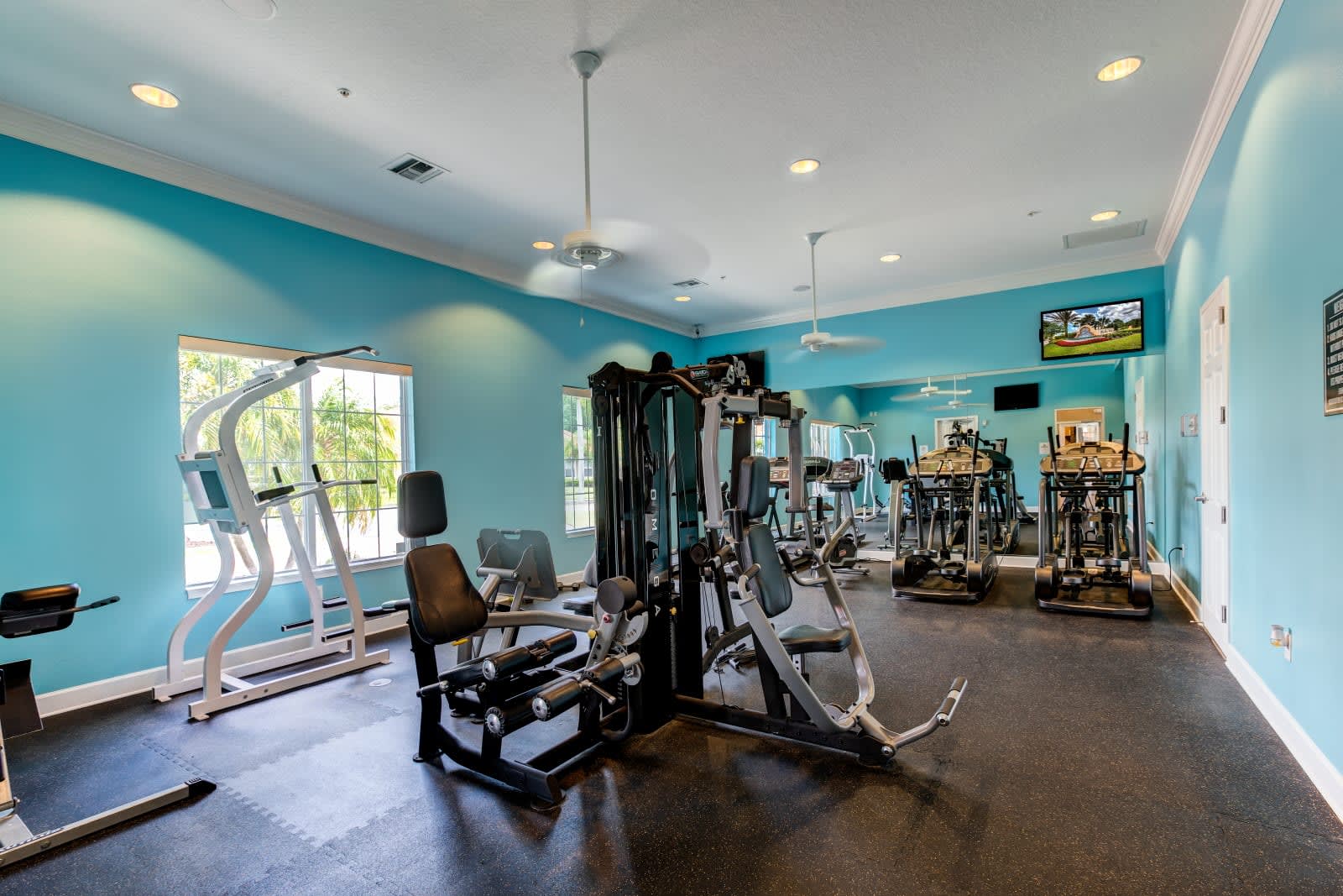 A well-equipped fitness center with light blue walls and black rubber flooring, featuring various exercise machines including treadmills, ellipticals, and weight machines. Large windows offer natural light, and ceiling fans provide air circulation.