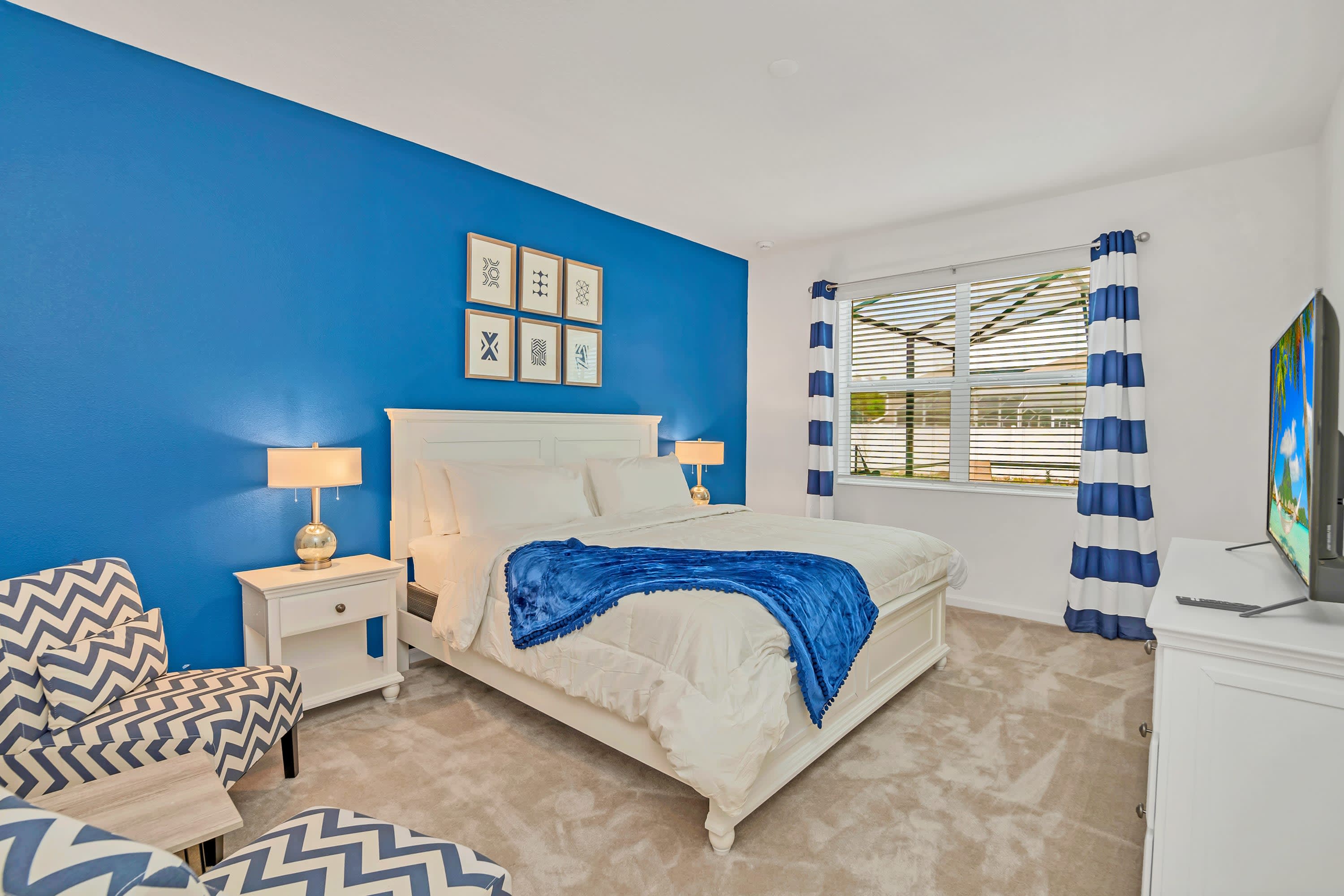 Bright bedroom with a bold blue accent wall behind a white queen bed, flanked by white nightstands with lamps, and a patterned blue and white armchair in the corner.