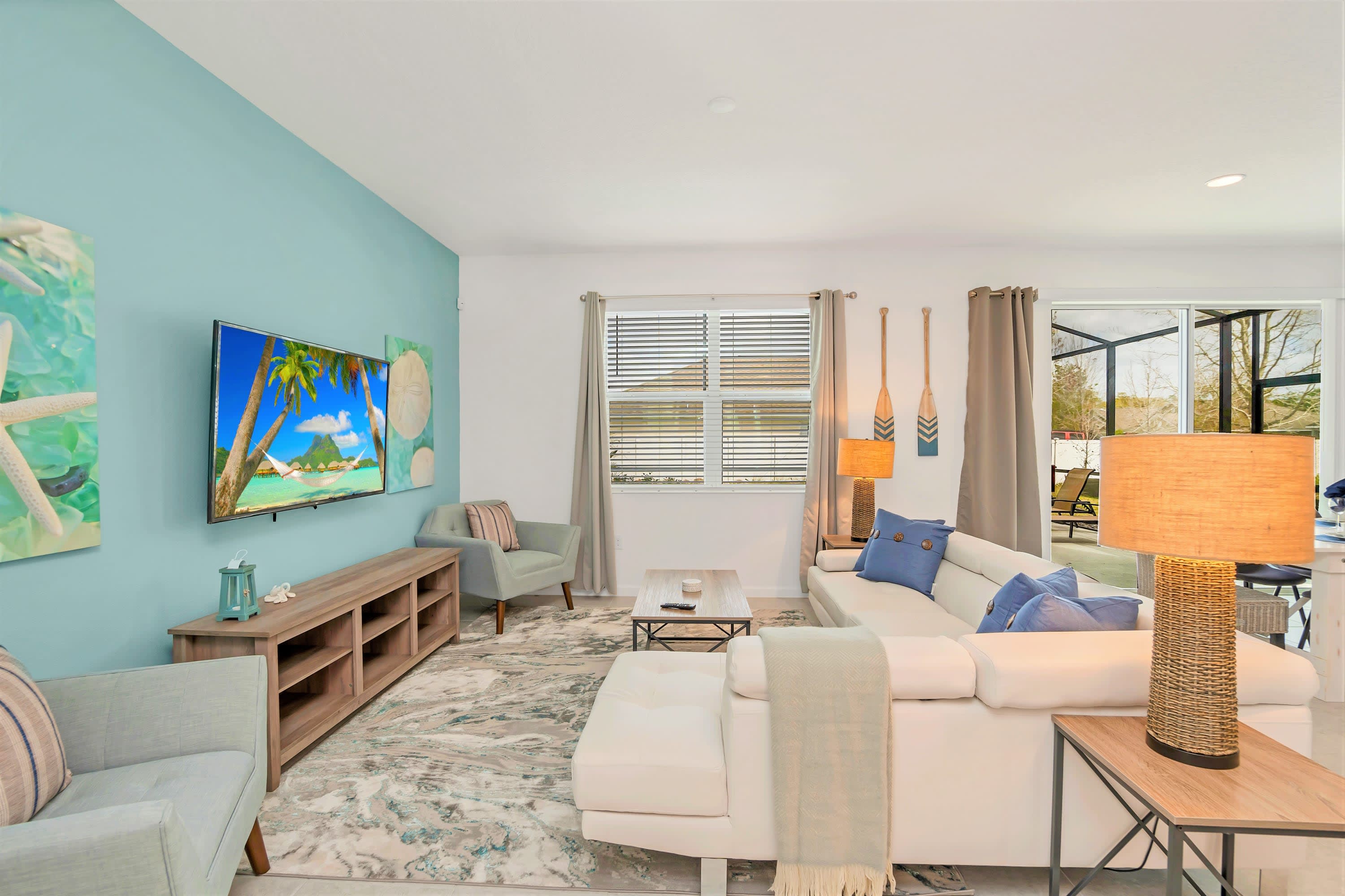 Bright living room with a white sectional sofa, wooden coffee table, and a wall-mounted TV, complemented by coastal decor including decorative oars and a teal accent wall. Sliding doors lead to the outdoor pool area, enhancing the open feel.