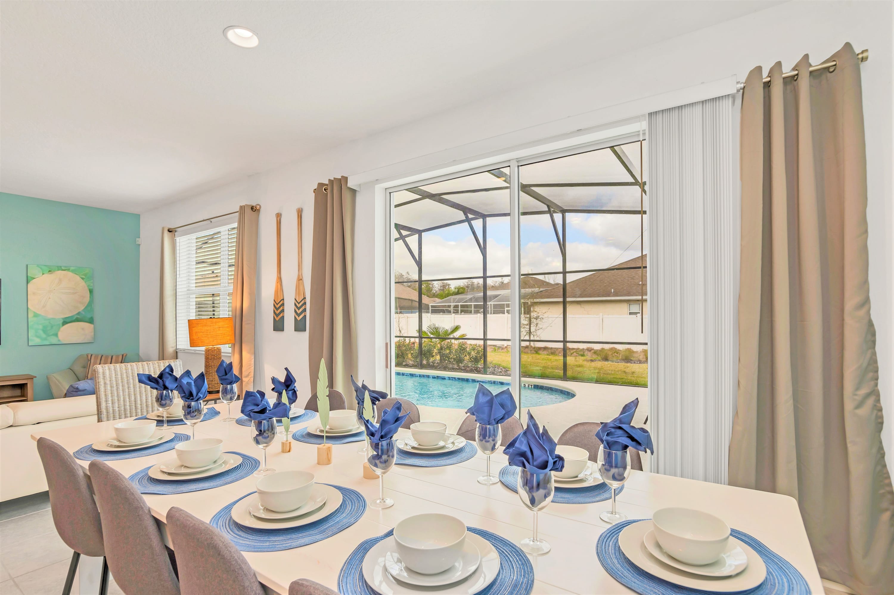 Dining area with a long white table elegantly set for eight, featuring blue place settings, and large sliding glass doors offering a direct view of the private screened pool. The living room is visible in the background, creating an open-concept flow.