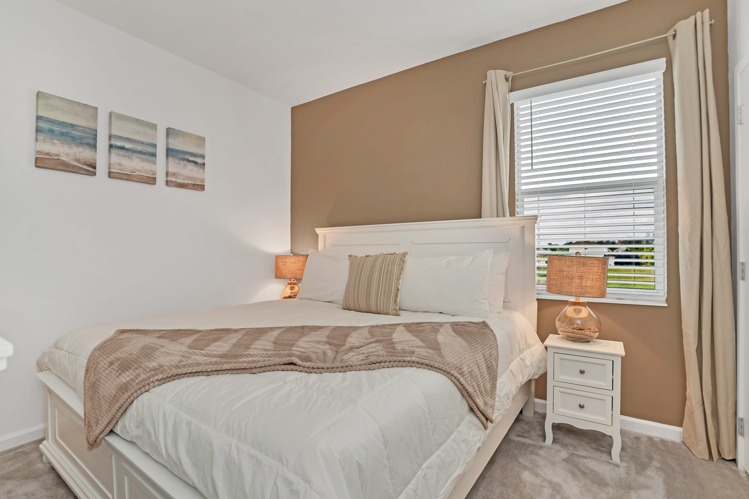 A bright bedroom featuring a white king-sized bed with a beige throw, two nightstands with lamps, and a warm brown accent wall. Three framed beach-themed artworks adorn the white wall, complementing the neutral decor.