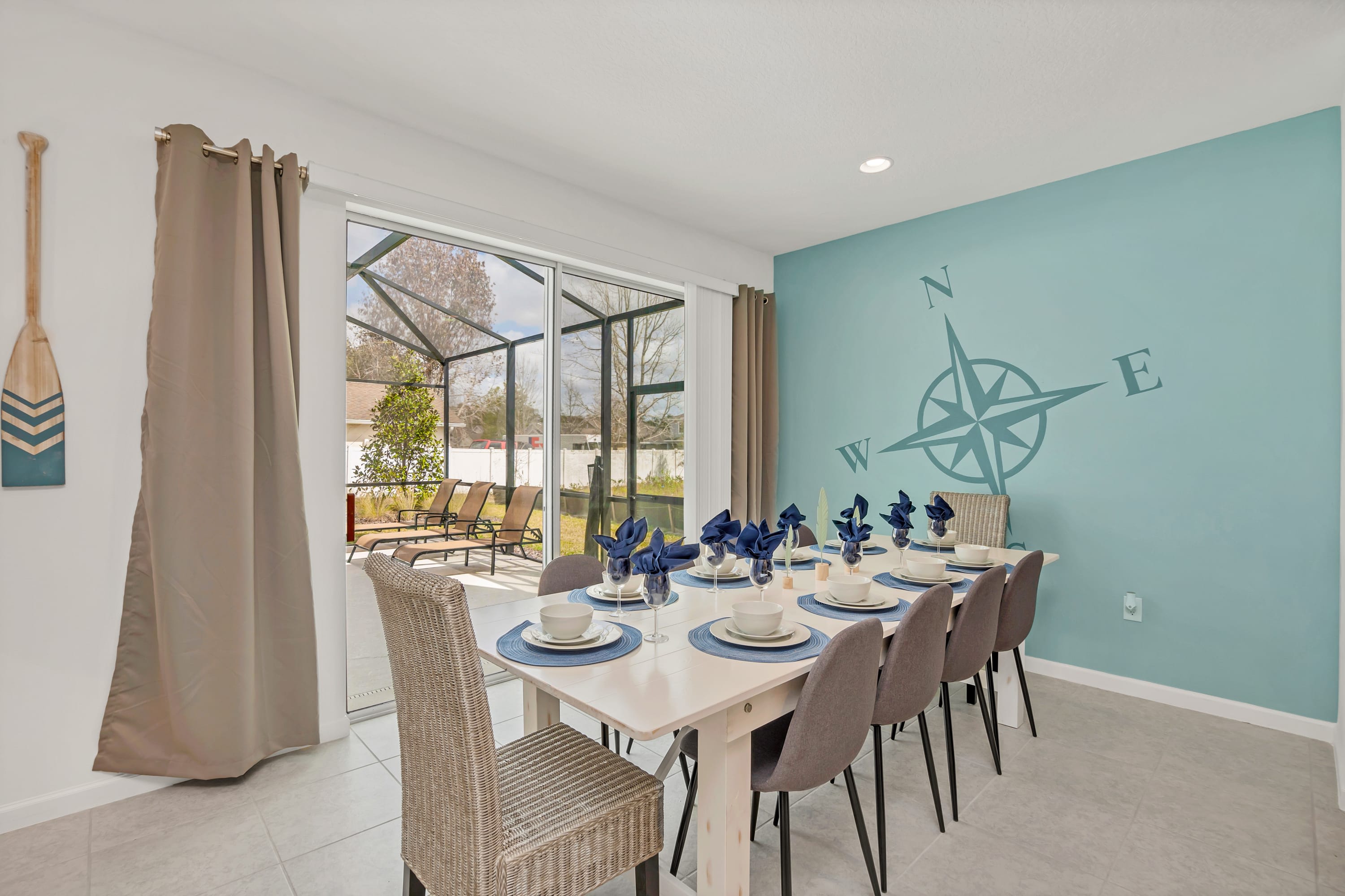 Coastal-themed dining room featuring a long white table set for eight with blue placemats and napkins, against a light teal accent wall adorned with a large compass rose decal. Sliding glass doors provide access to the screened pool area.