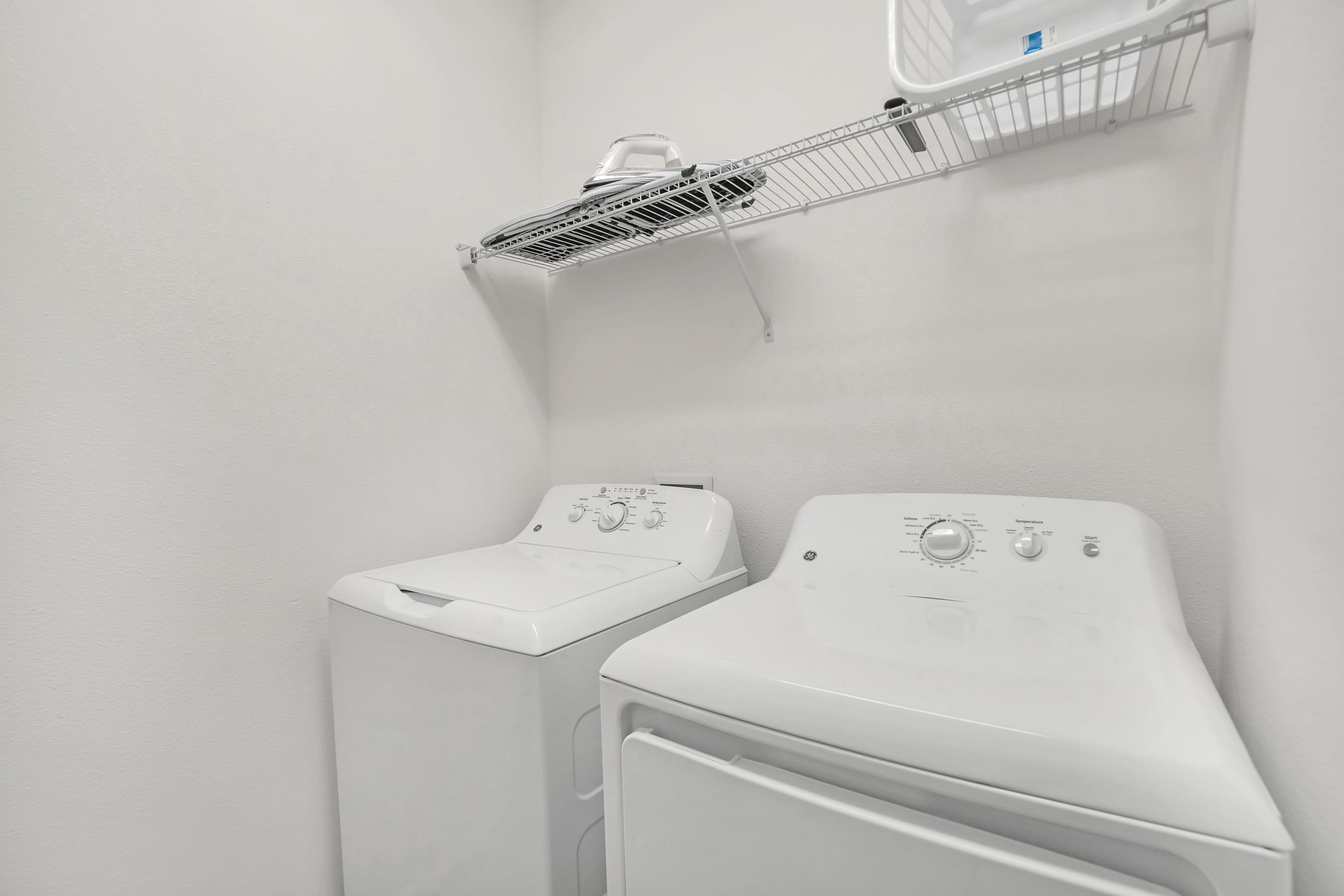 A functional laundry room equipped with a white top-loading washing machine and a front-loading dryer. A wire shelf with an ironing board and iron is mounted above the appliances for convenience.