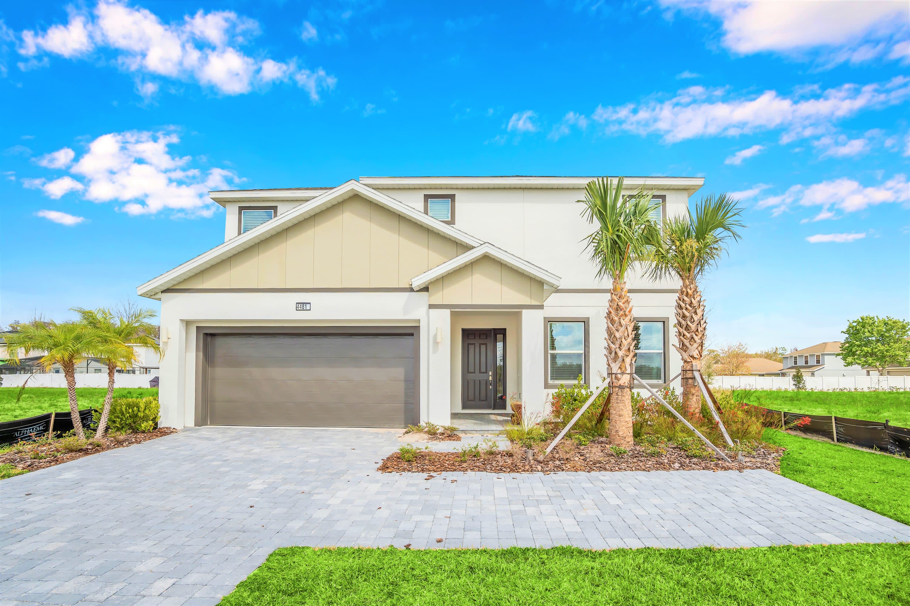 A modern two-story house with a light-colored facade, a dark gray garage door, and a paved driveway. Palm trees are planted in the front yard, and the sky is bright blue with white clouds.