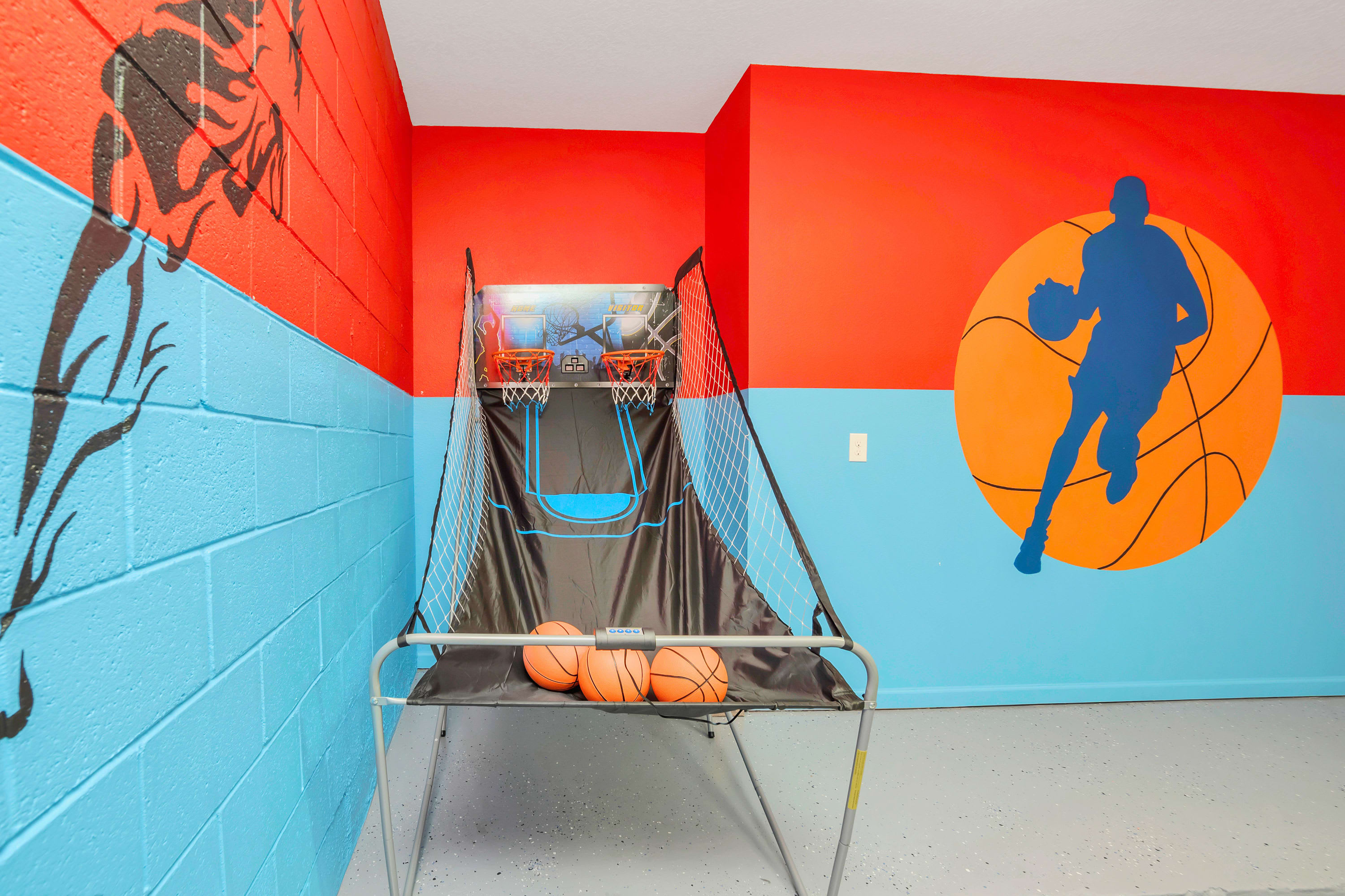 Vibrant game room with a basketball arcade game, featuring two hoops and three basketballs, set against a backdrop of red and blue painted walls with sports-themed murals.