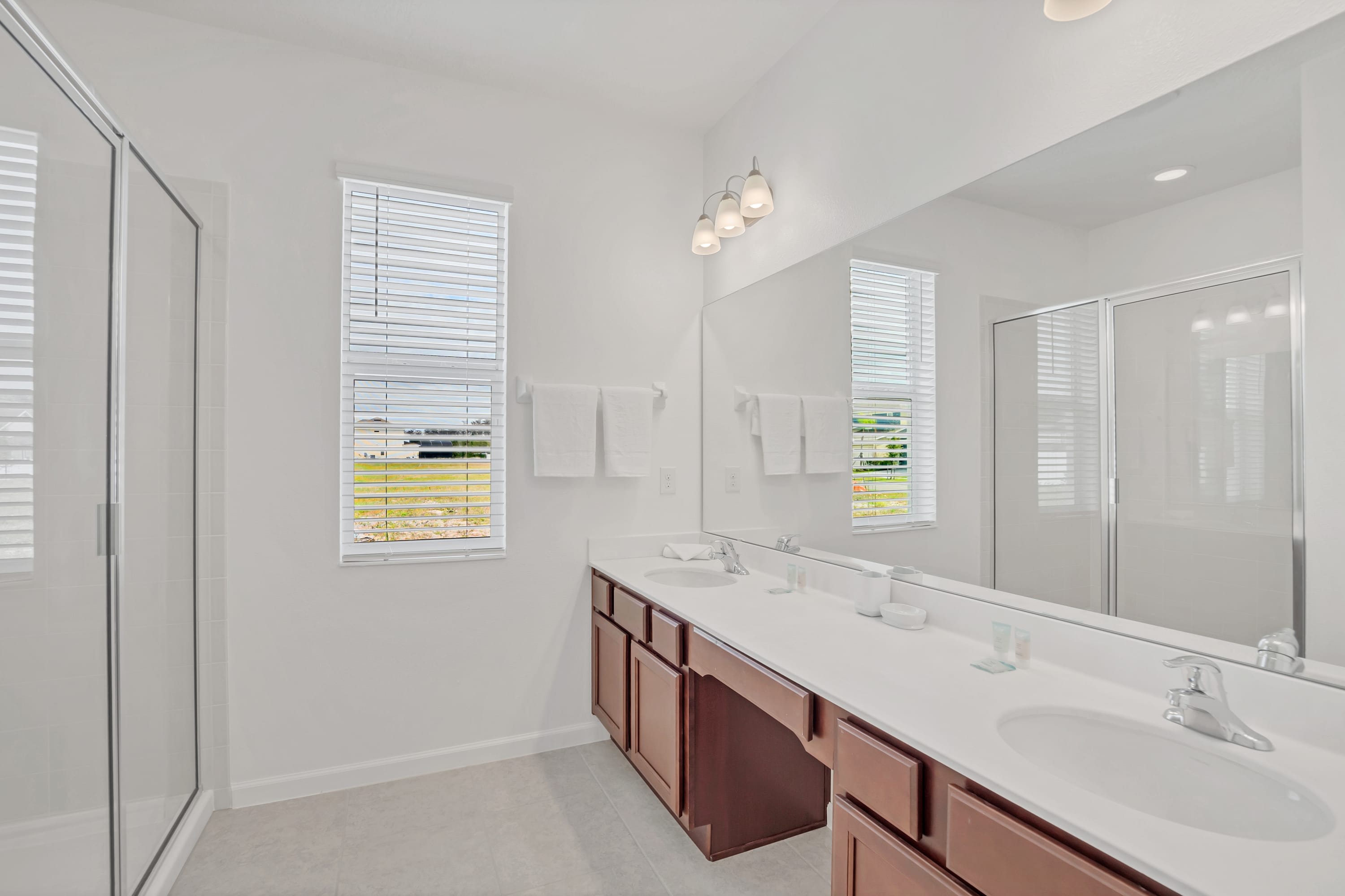 Spacious white bathroom with a long double vanity featuring dark wood cabinets and white countertops, a large mirror, and a glass-enclosed shower.