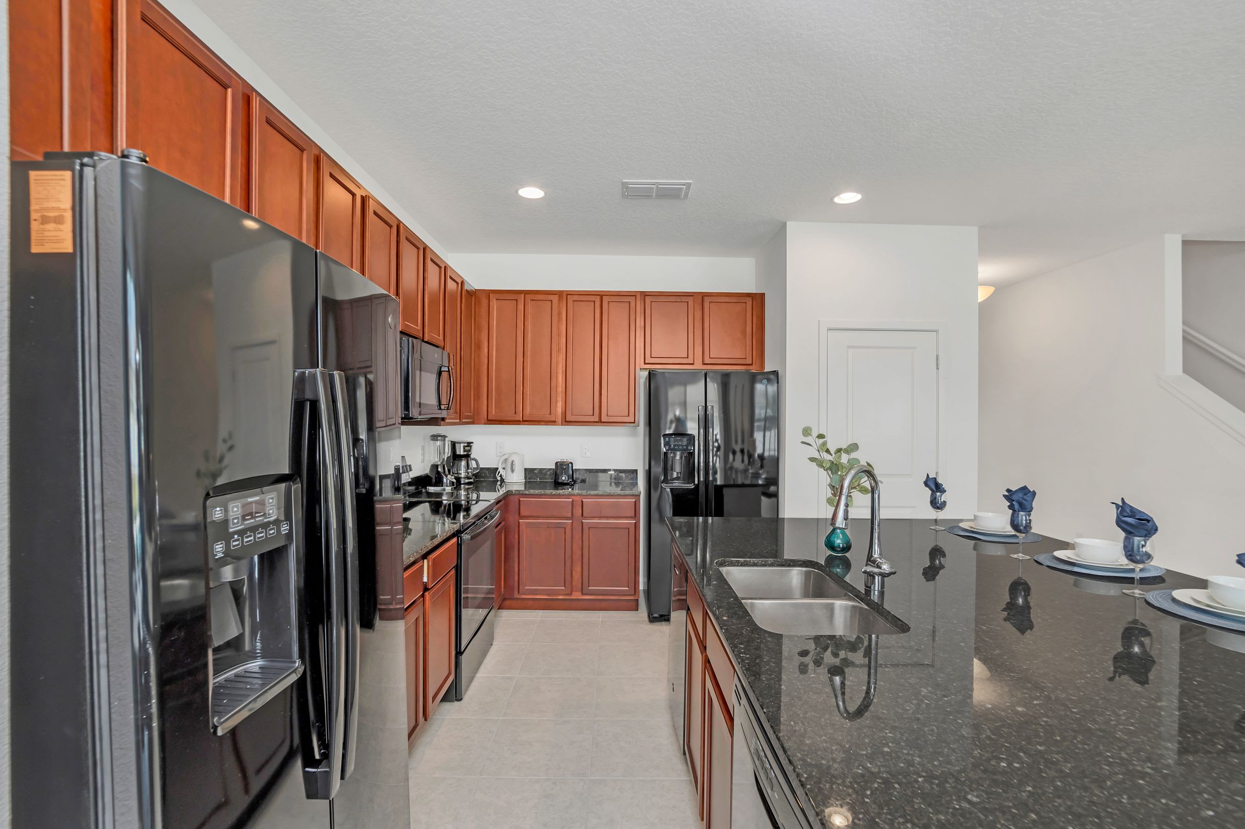 Modern kitchen featuring dark granite countertops, rich cherry wood cabinets, and sleek black appliances, including two refrigerators and a large island with an undermount sink.