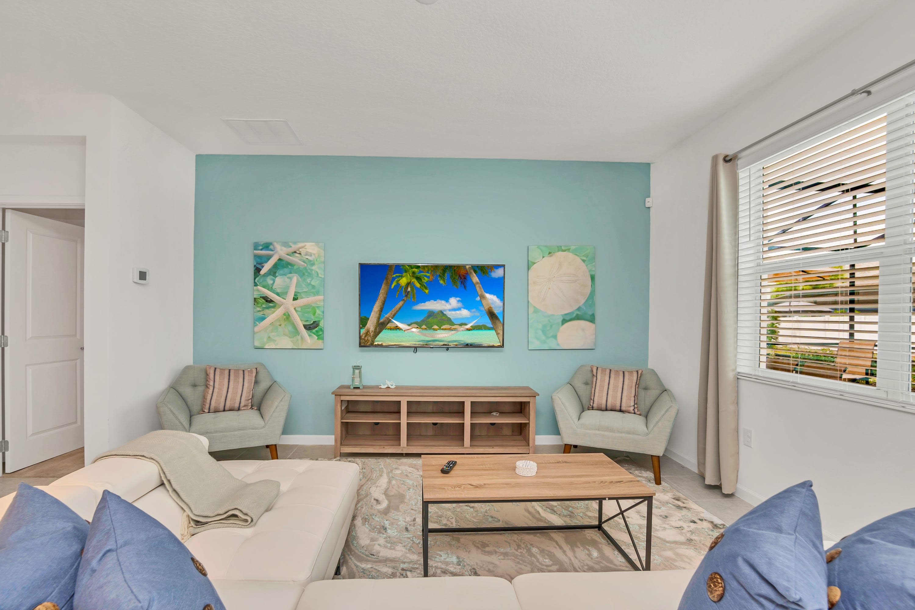 Cozy living room featuring a large wall-mounted TV above a wooden console, flanked by two armchairs and coastal-themed artwork on a light teal accent wall. A white sectional sofa and a patterned area rug complete the inviting space.
