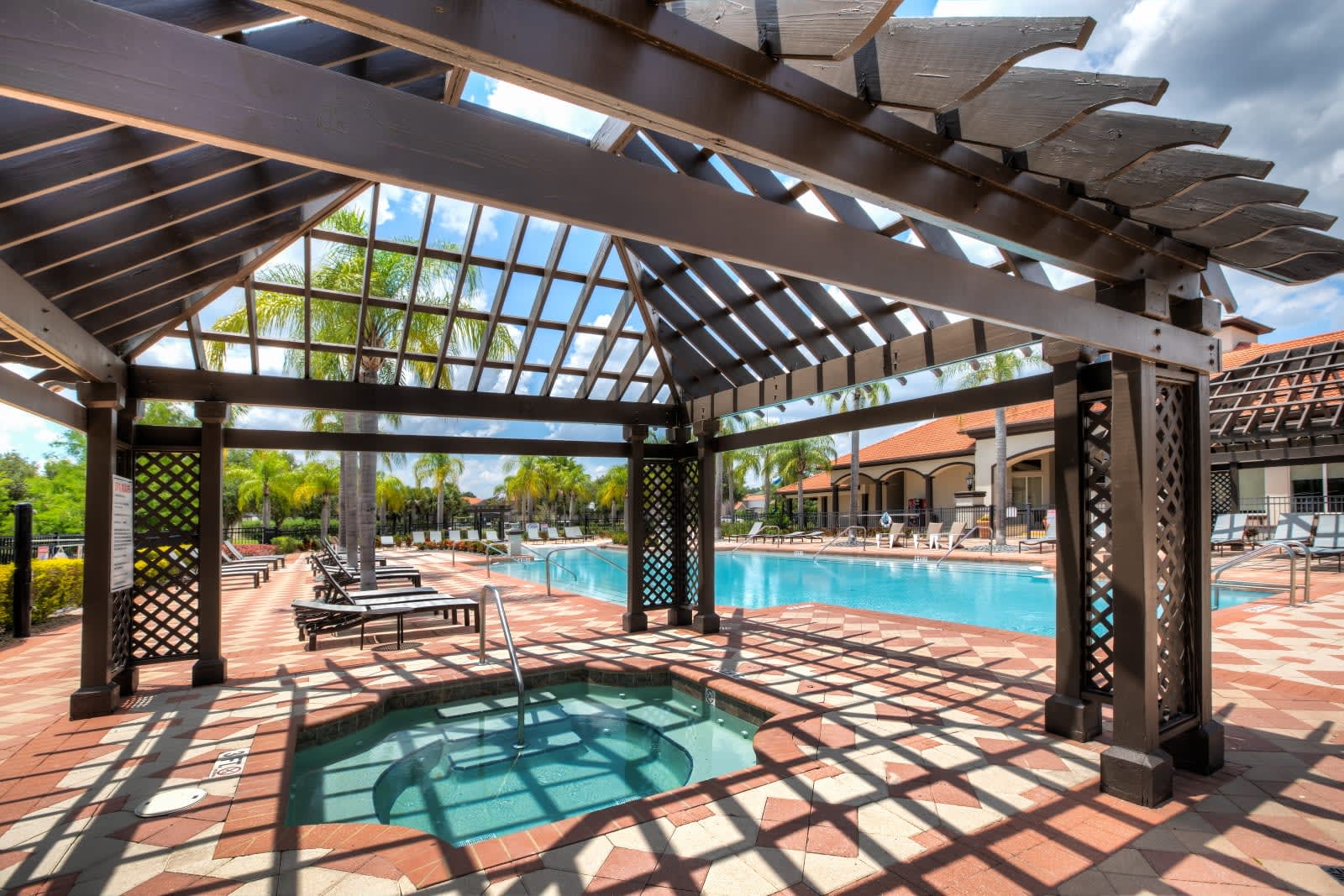 A close-up view of a small, square hot tub with steps, located under a large dark wooden pergola with slatted roof. The hot tub is surrounded by patterned brick pavers, with lounge chairs and a swimming pool visible in the background.