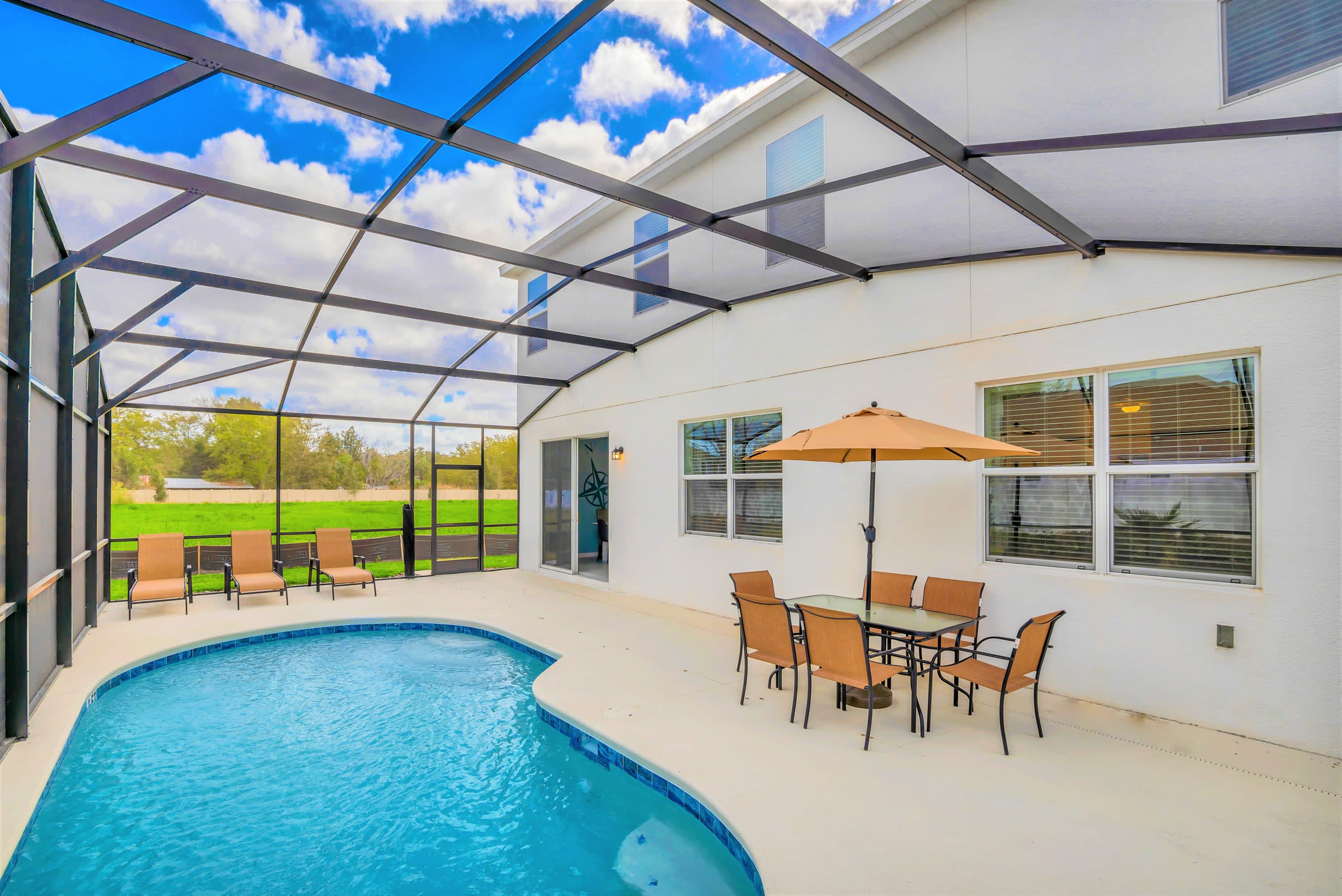 Expansive screened-in pool deck with a private swimming pool, multiple lounge chairs, and an outdoor dining set with an umbrella, all set against the white exterior of the house. The enclosure provides views of the surrounding landscape.