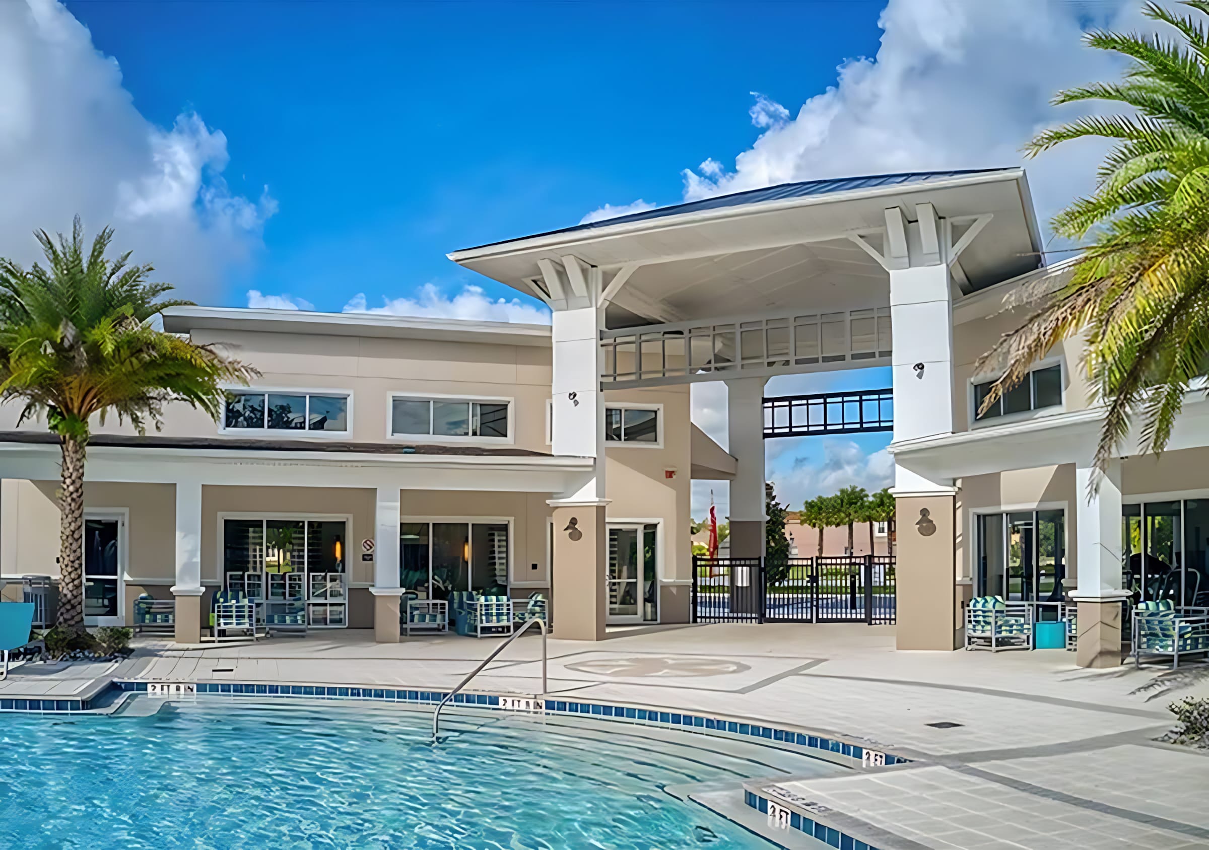 Expansive communal pool area with a large swimming pool, surrounded by lounge chairs and palm trees. A modern clubhouse building with a covered entrance stands in the background under a blue sky.