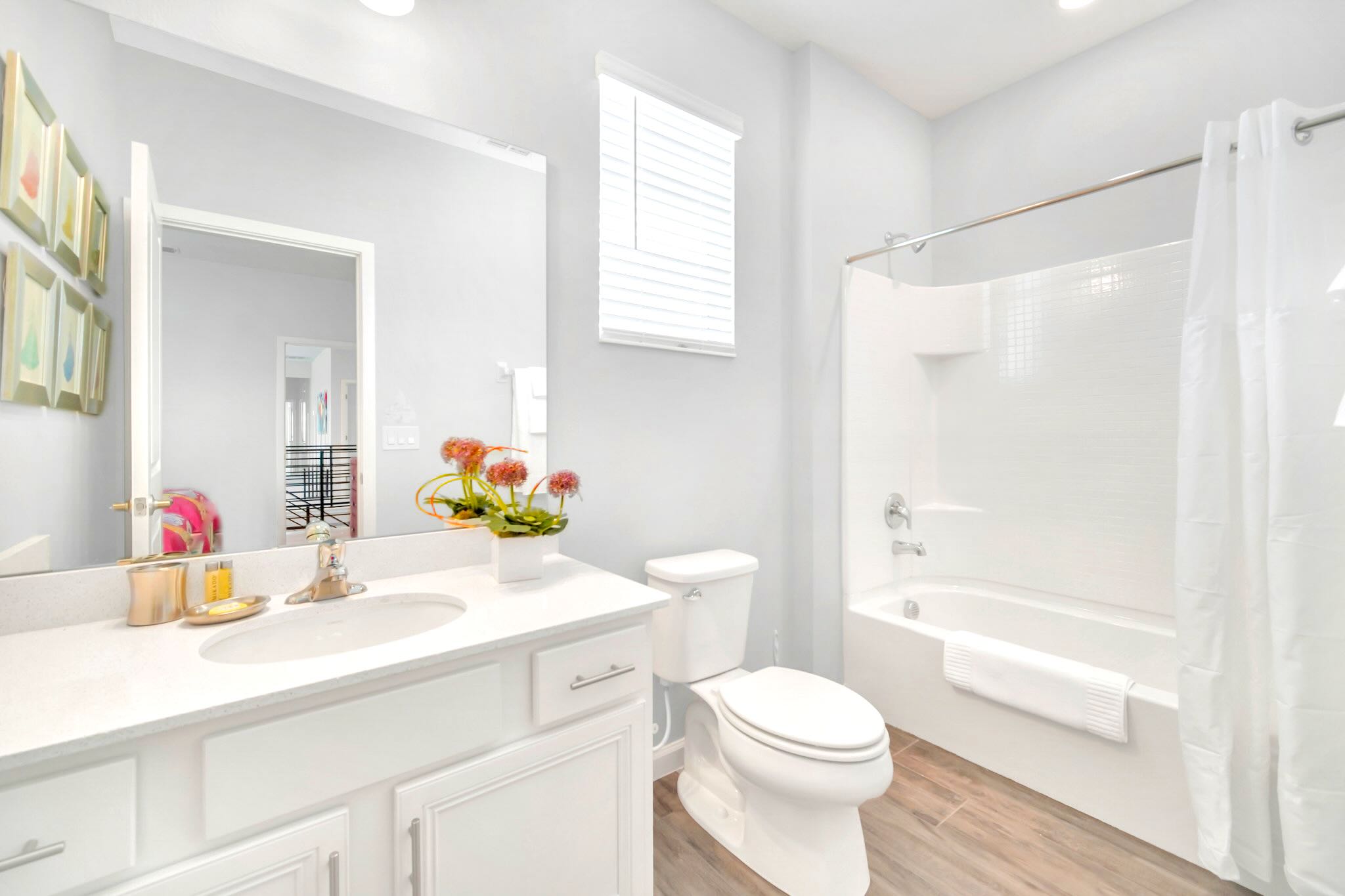 A clean and bright bathroom with a white single vanity, a large mirror, and a toilet. It features a white bathtub with a shower curtain and light wood-look tile flooring, complemented by a window with blinds.