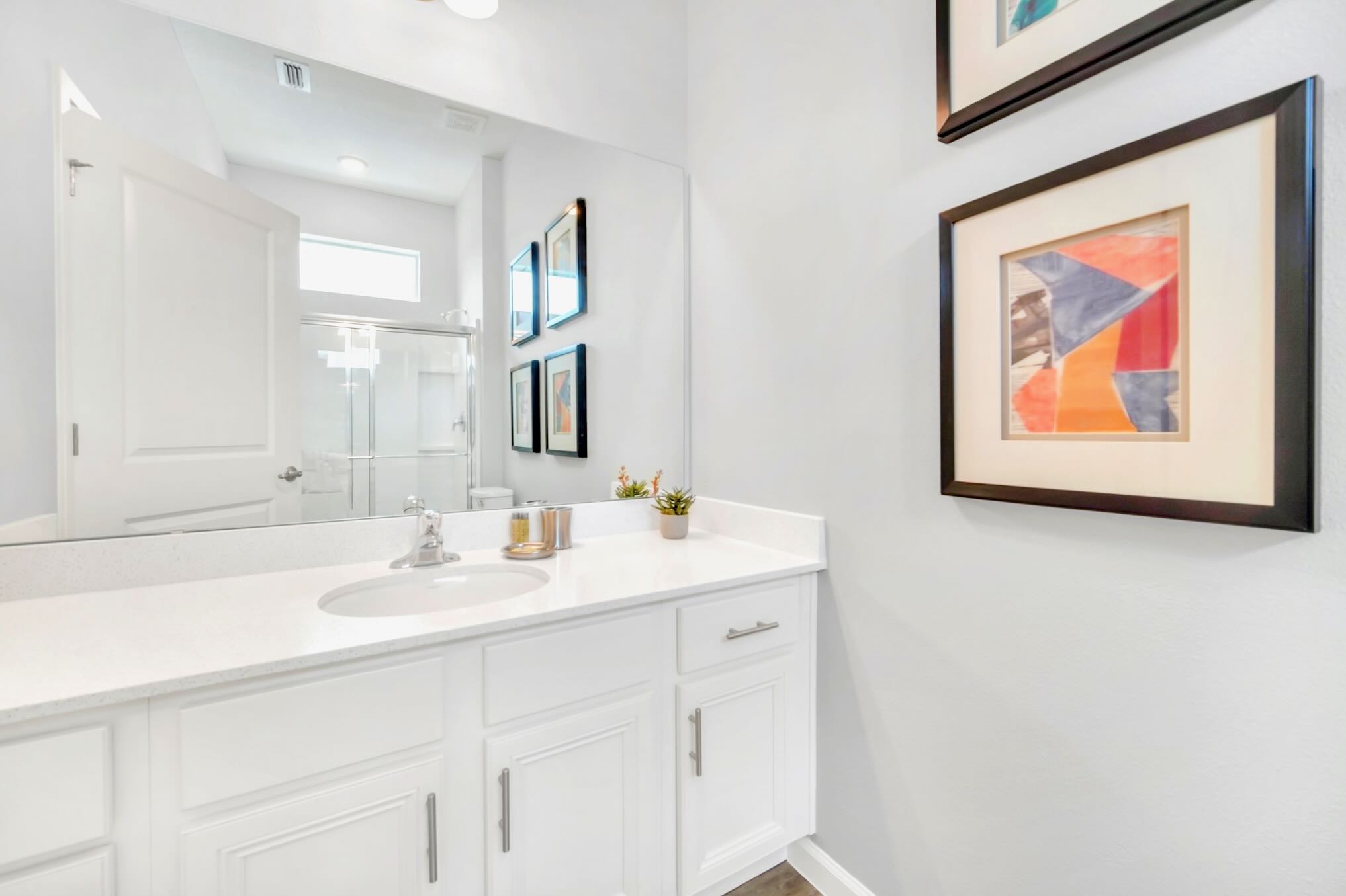 A modern bathroom with a white single vanity, a large mirror, and a white quartz countertop. The light gray walls are adorned with framed abstract artwork, and a glass-enclosed shower is visible in the mirror's reflection.