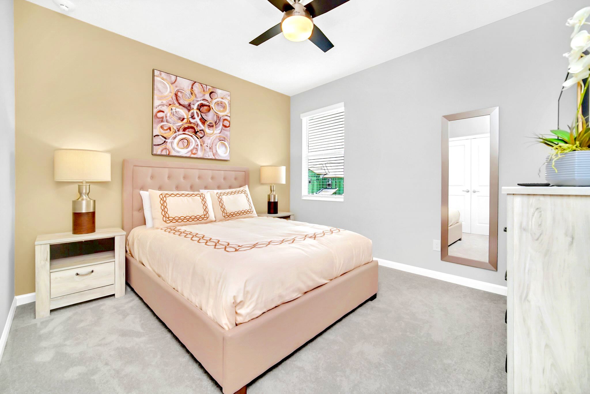 A stylish bedroom with a queen-sized bed featuring a light pink upholstered headboard and matching bedding with elegant embroidered details. The room has a beige accent wall, light gray side walls, two nightstands with lamps, and a full-length mirror.