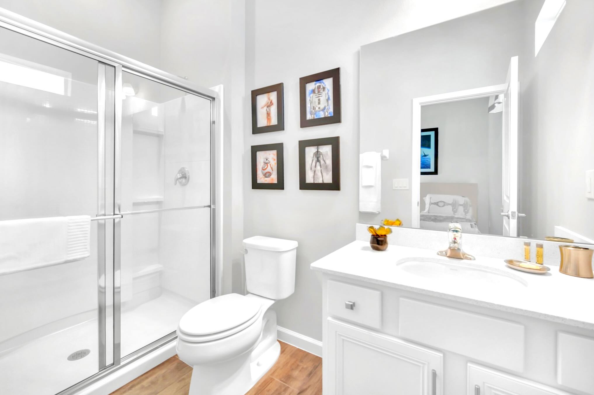 A modern bathroom with a white single vanity, a large mirror, and a toilet, featuring a glass-enclosed walk-in shower. The light gray walls are decorated with framed Star Wars artwork, and the floor has light wood-look tile.