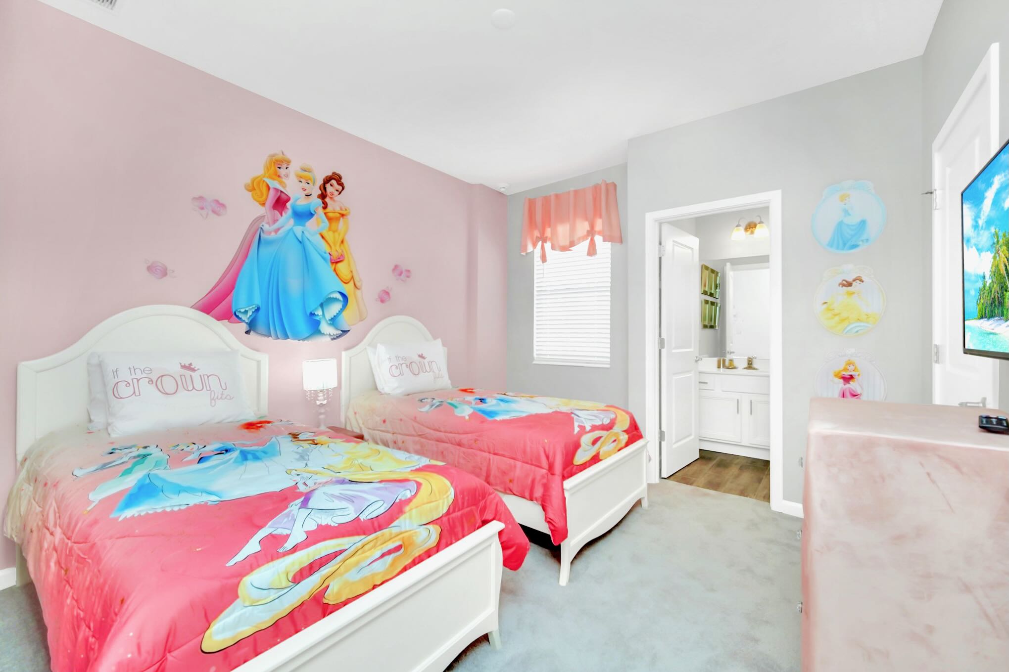 A vibrant children's bedroom with two white twin beds, each adorned with princess-themed comforters and wall decals of Disney princesses. The room features a pink accent wall, light gray side walls, a window with a pink valance, and a TV on a dresser.