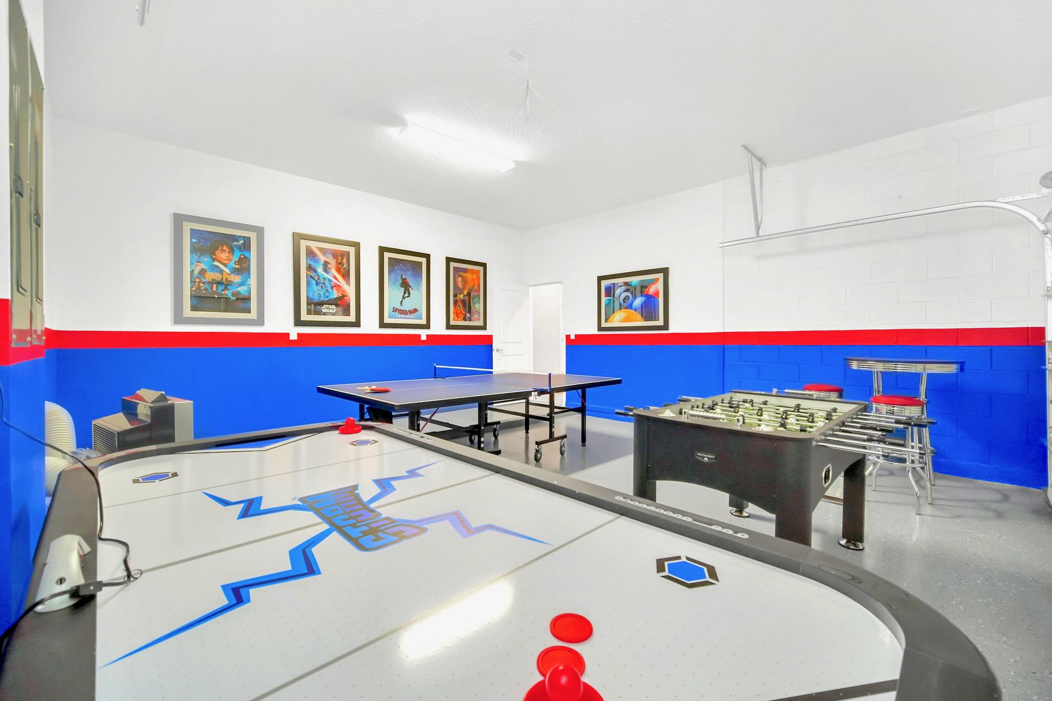 A vibrant game room, likely a converted garage, features an air hockey table, a ping pong table, and a foosball table, set against walls painted with blue and red accents.