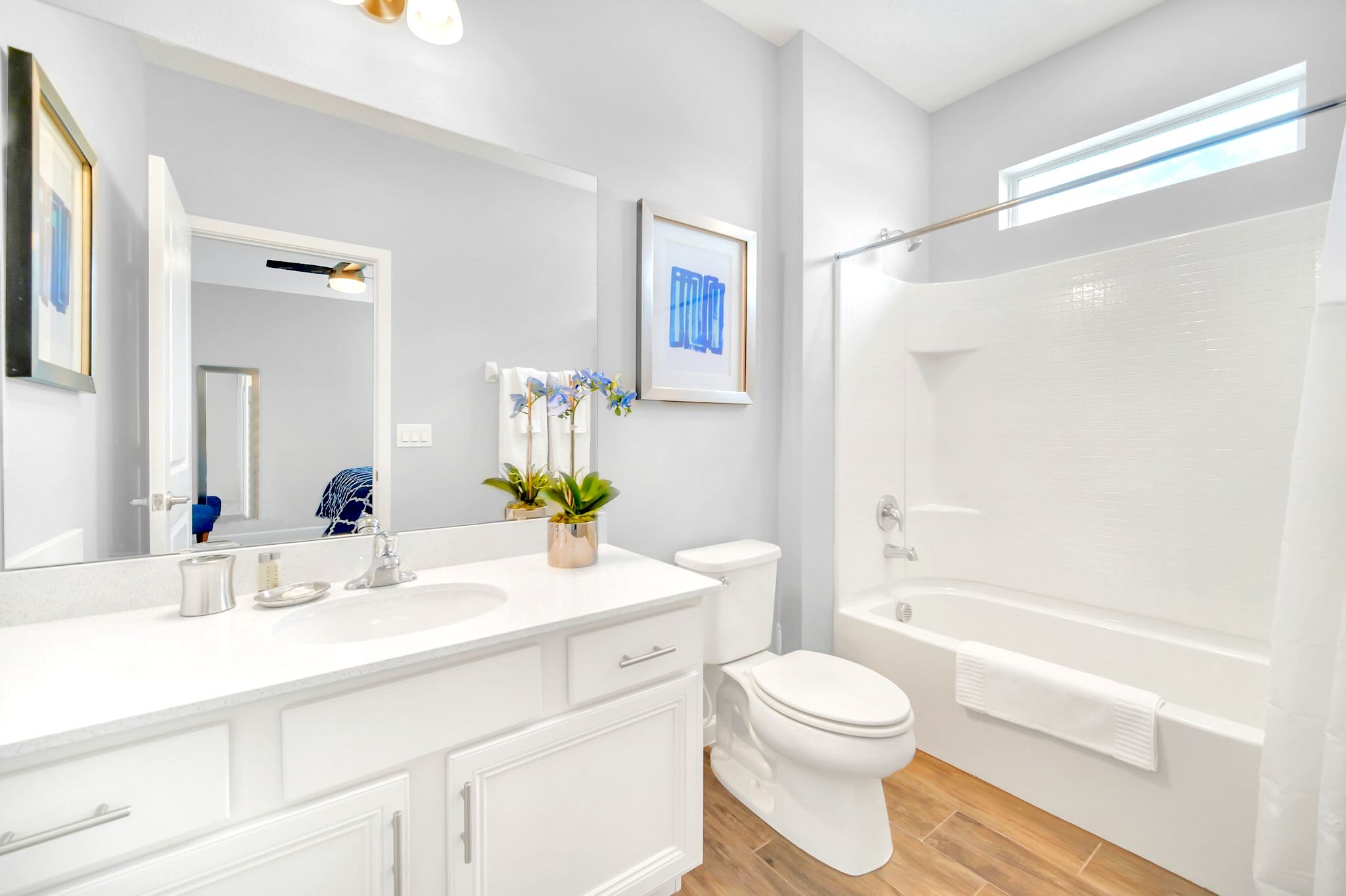 A bright bathroom featuring a single white vanity, a large mirror, and a bathtub with a shower. Wood-look tile flooring and light gray walls create a clean and modern space.