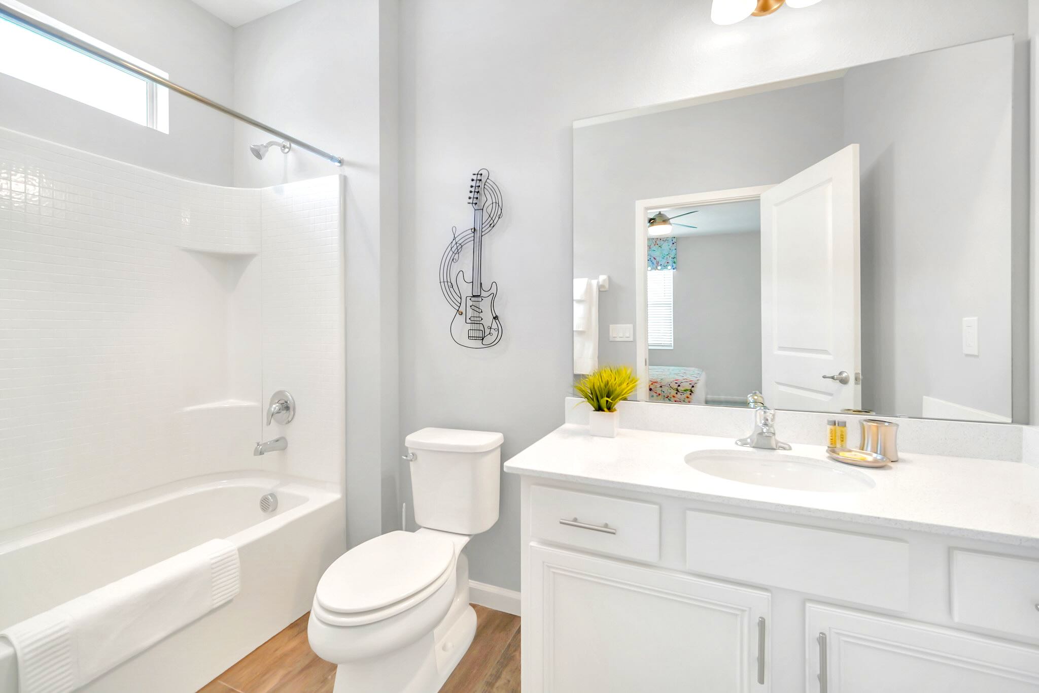 A clean bathroom with a single white vanity, a large mirror, and a bathtub with a shower. Wood-look tile flooring and a guitar-themed wall art add a touch of personality.