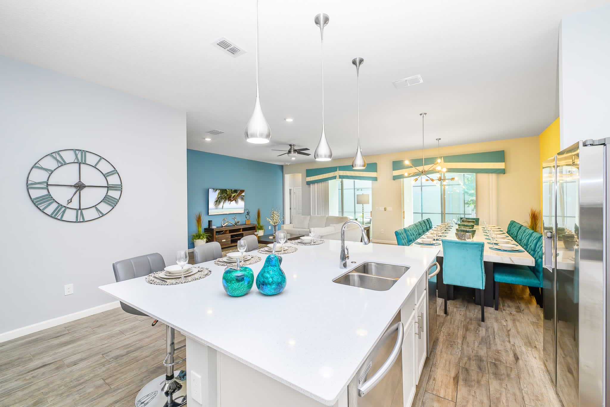 View from the kitchen island, showcasing its white quartz countertop and bar seating, looking towards the open-concept living and dining areas. The living room features a blue accent wall and white sofas, while the dining area has teal chairs.