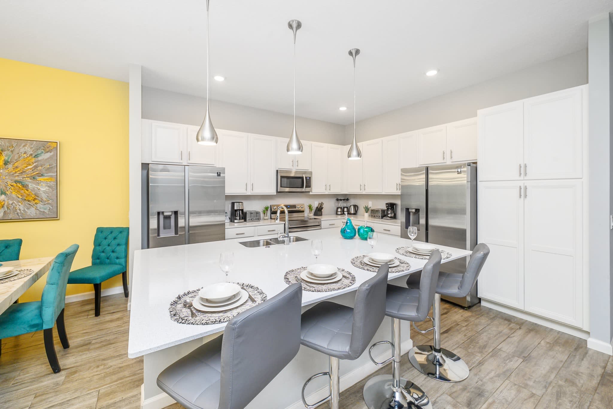 Modern kitchen with white shaker cabinets, stainless steel appliances, and a large white quartz island featuring a sink and three grey bar stools. Contemporary pendant lights hang above the island, and a vibrant yellow wall is visible to the left.