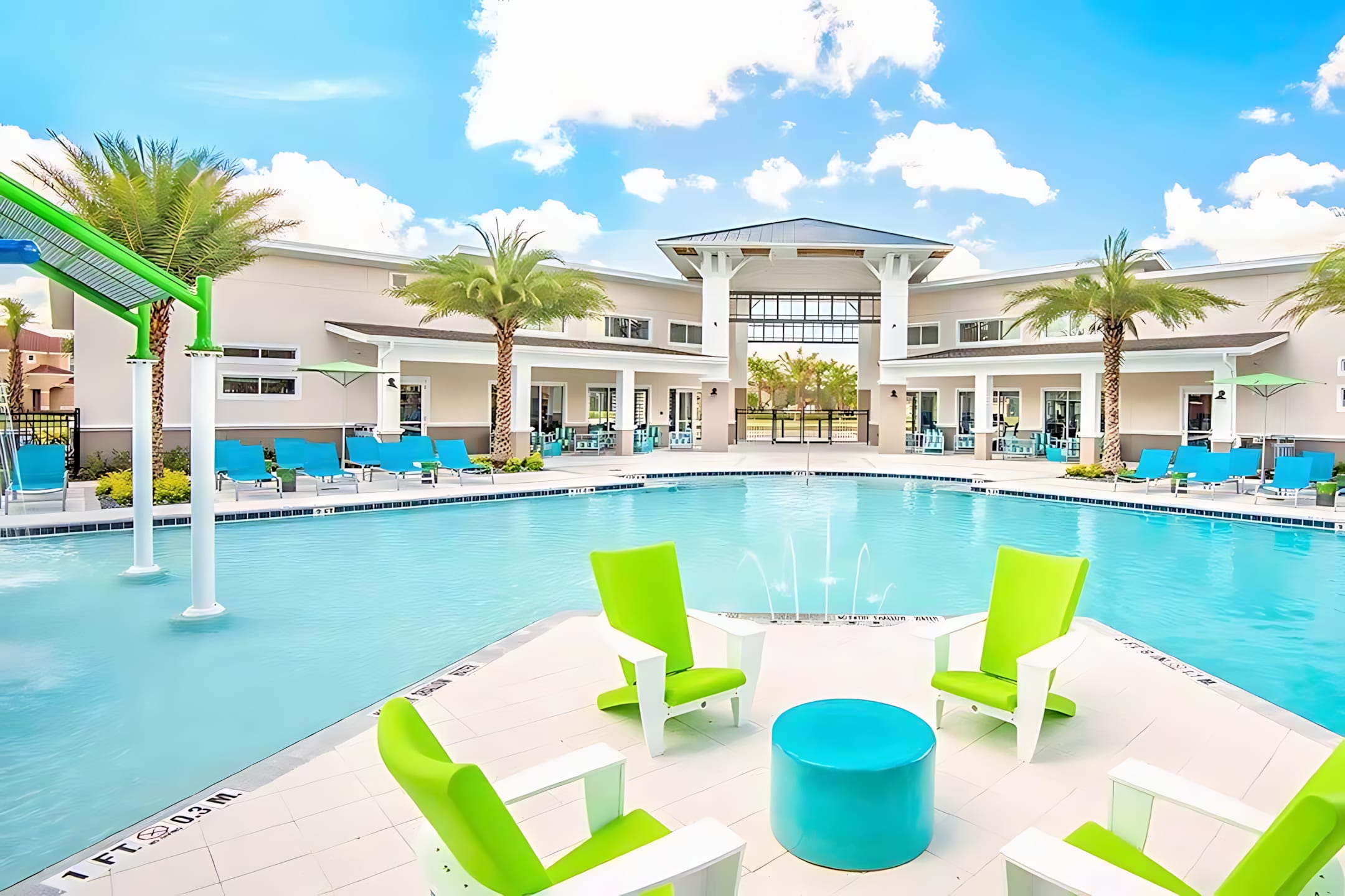 Vibrant communal pool area with a large, irregularly shaped pool featuring a shallow entry, water fountains, and bright green and blue lounge chairs. A modern clubhouse is visible in the background.