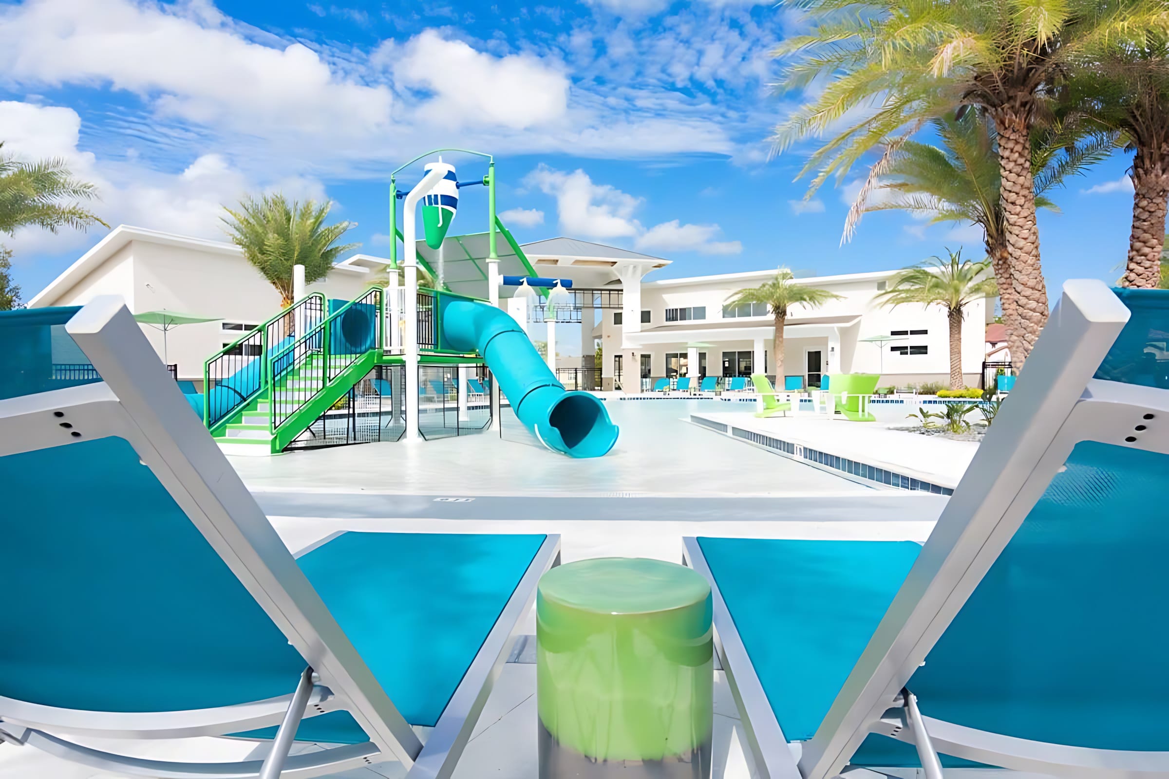 A wide shot of the communal pool area shows a shallow water play zone with a blue and green slide and water features, surrounded by numerous blue lounge chairs. Palm trees and a contemporary white building with large windows are visible under a bright sky.