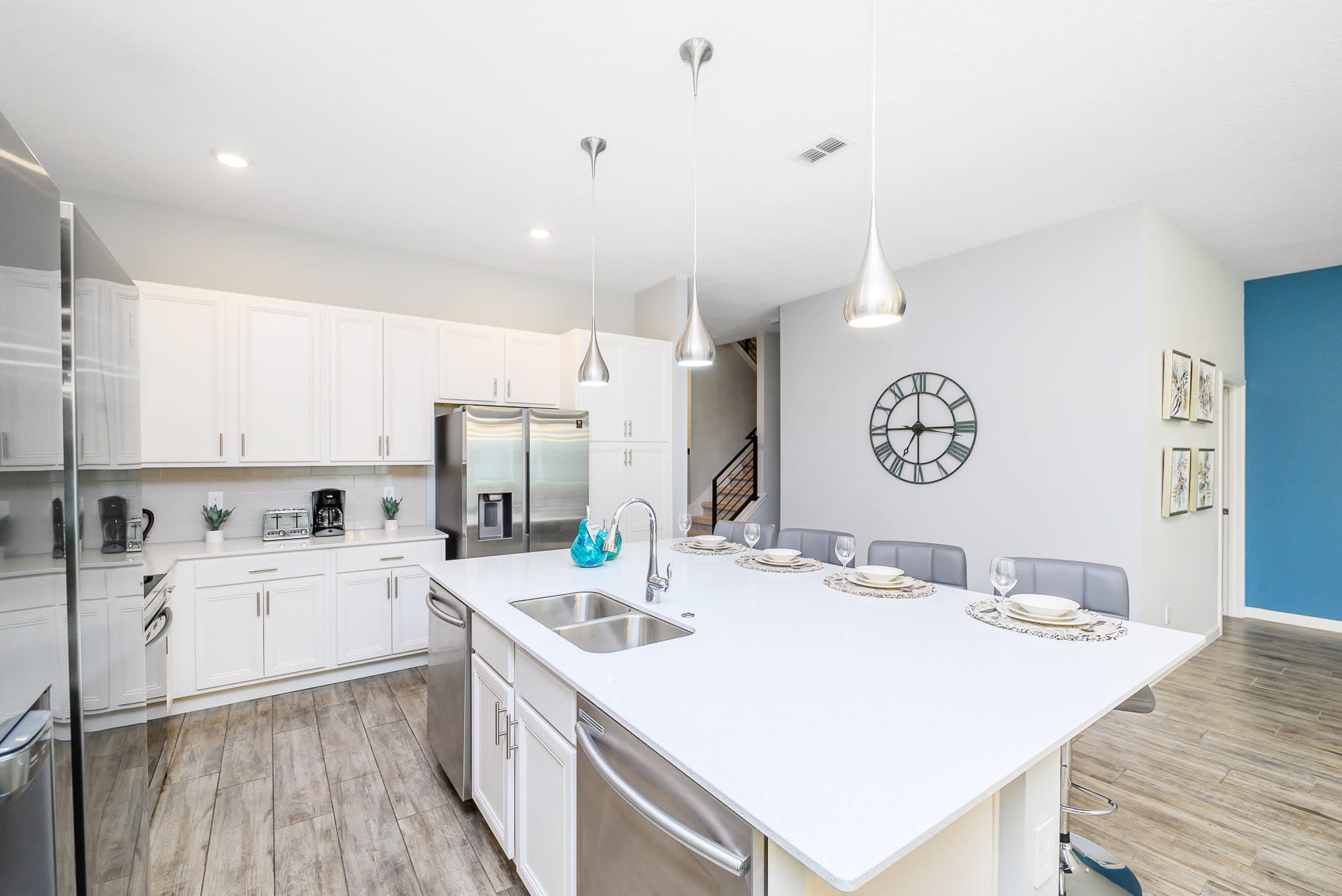 Spacious white kitchen featuring extensive cabinetry, stainless steel appliances, and a large central island with a sink and seating for four. Modern pendant lights illuminate the island, and a large decorative wall clock is visible in the adjacent hallway.