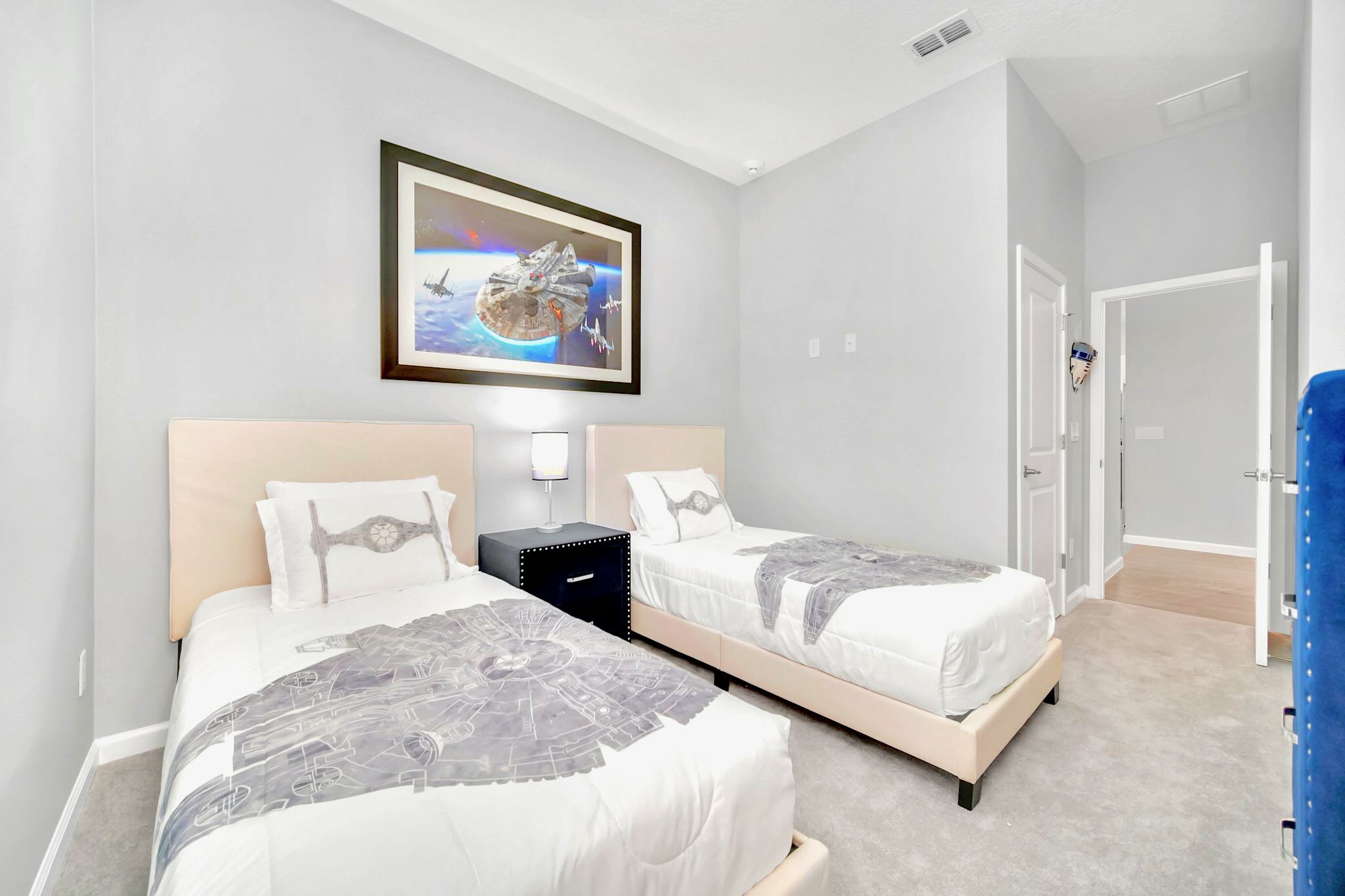 A children's bedroom designed with a Star Wars theme, featuring two twin beds with light upholstered headboards and themed bedding. A large framed Star Wars artwork hangs above the beds, complemented by a black nightstand and lamp in between.