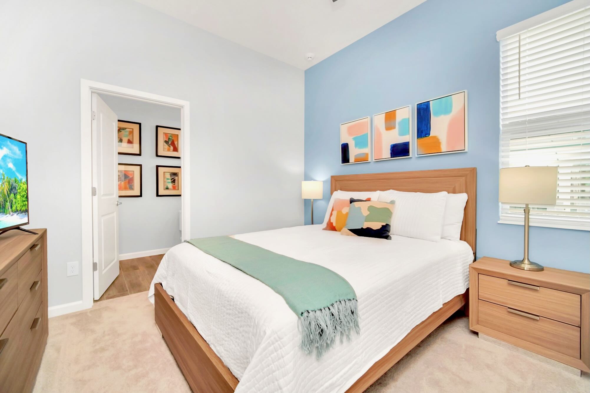 A bright bedroom featuring a queen-sized bed with a wooden headboard, white and light blue bedding, and a light green throw. The room has a light blue accent wall, light-colored carpet, and a wooden nightstand with a lamp, plus a TV on a matching dresser.