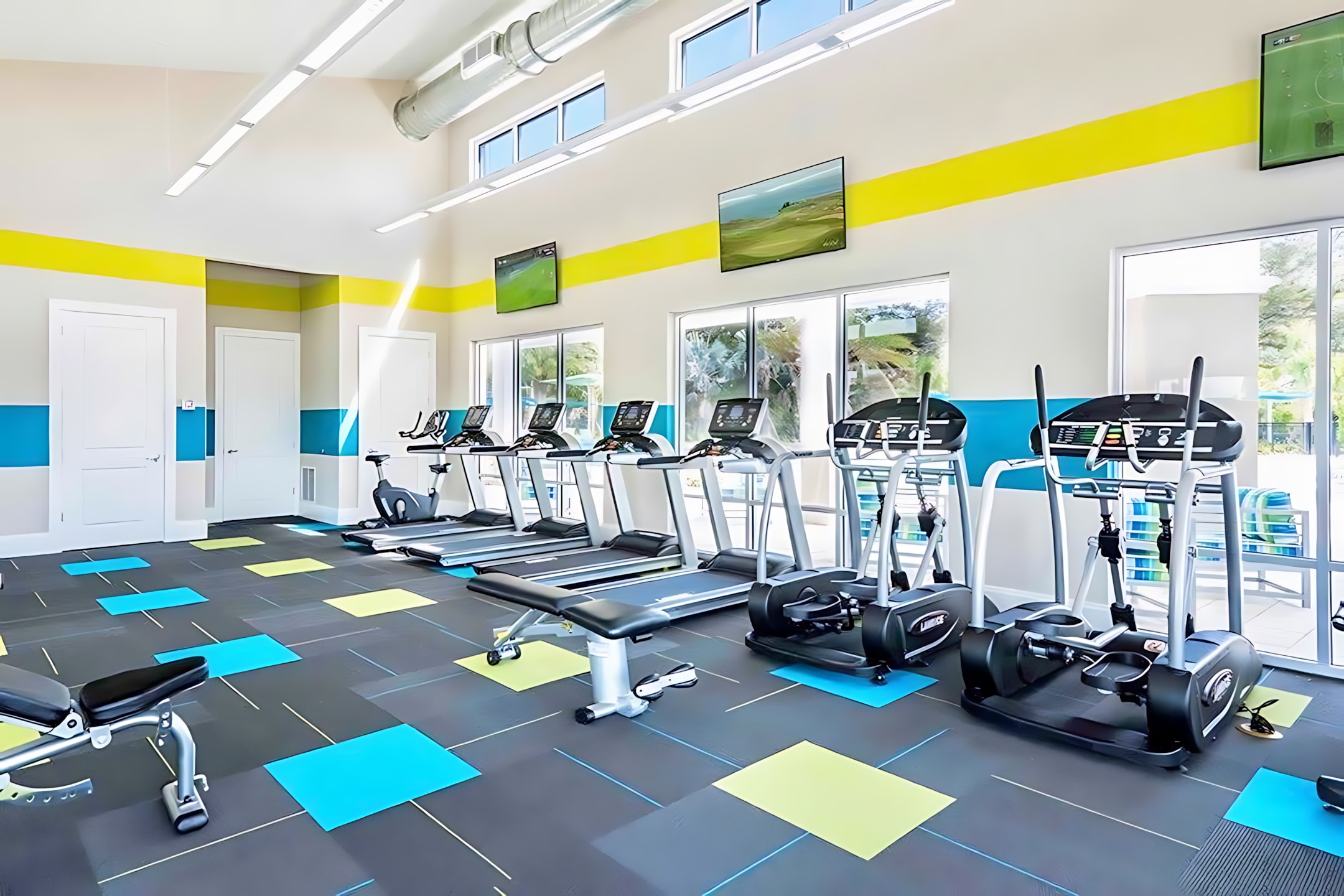 A spacious, well-lit fitness center features multiple treadmills, elliptical machines, and weight benches on a dark floor with blue and yellow square accents. Large windows overlook the outdoor pool area, and flat-screen TVs are mounted on the walls.