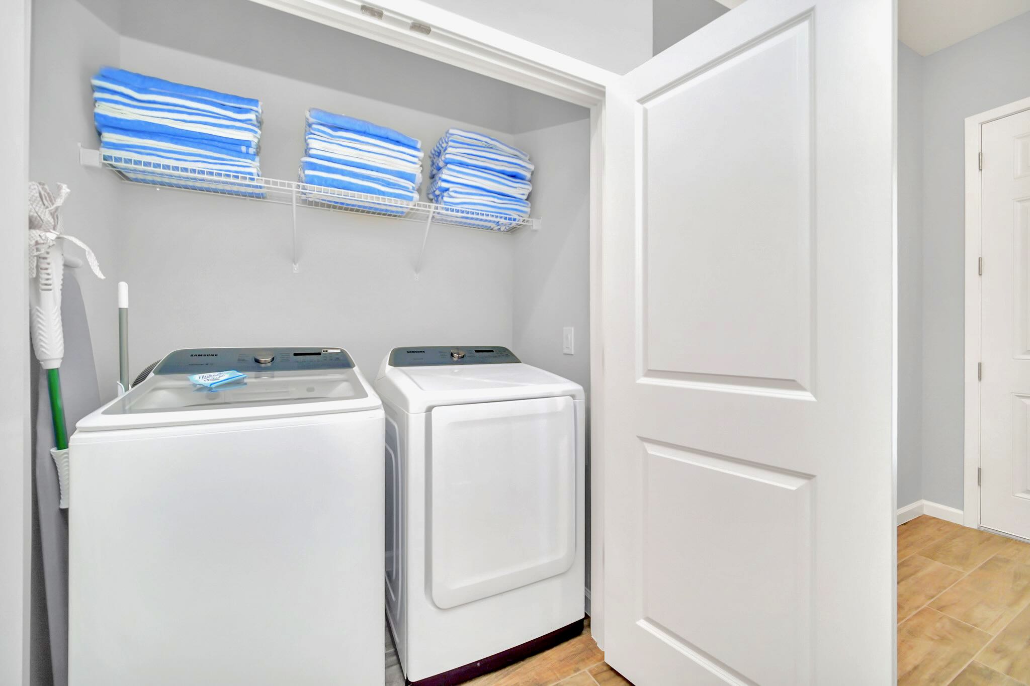 Compact laundry closet featuring a white top-load washing machine and a matching front-load dryer. A wire shelf above holds neatly folded blue and white striped towels.